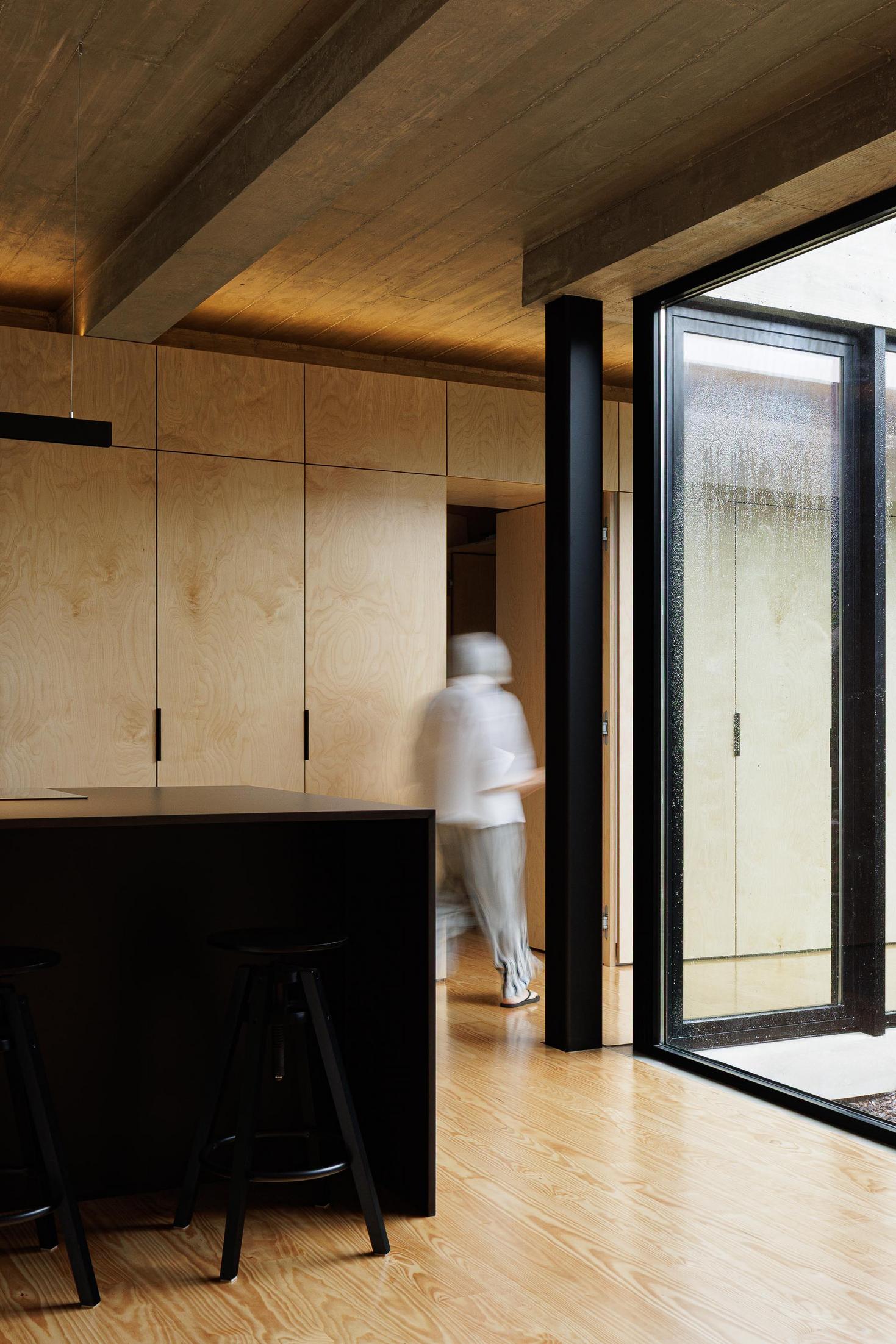 Cozinha: Ilha central preta, armários em contraplacado e pavimento em madeira. Porta envidraçada para pátio interior. — Casa CR arquitetura SO Arquitetura & Design em São Miguel