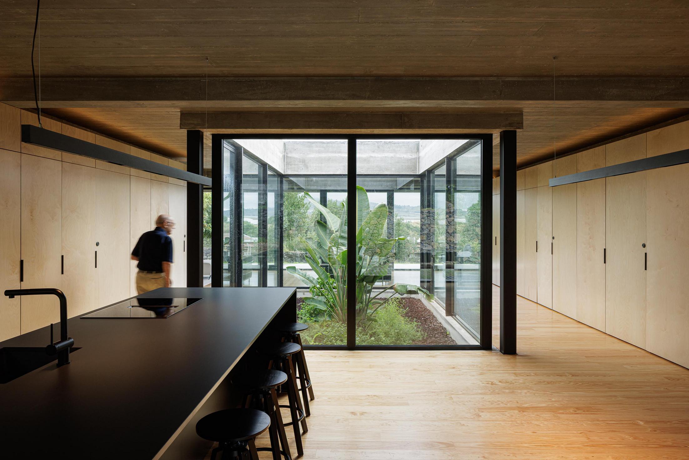 Cozinha: Ilha central preta, armários em contraplacado e pavimento em madeira. Pátio interior com vegetação. — Casa CR arquitetura SO Arquitetura & Design em São Miguel