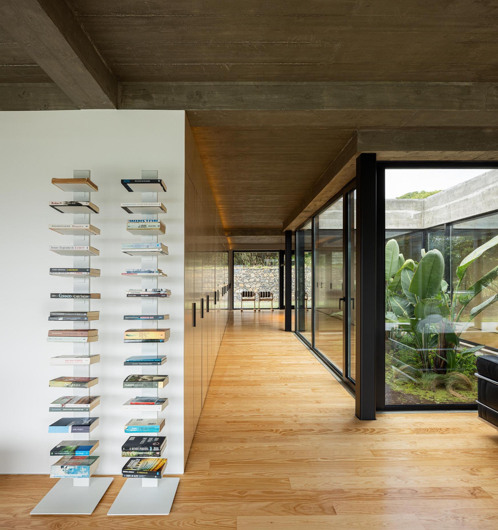Corredor: Estante com livros, armários em contraplacado e pavimento em madeira. Pátio interior com vegetação. — Casa CR arquitetura SO Arquitetura & Design em São Miguel