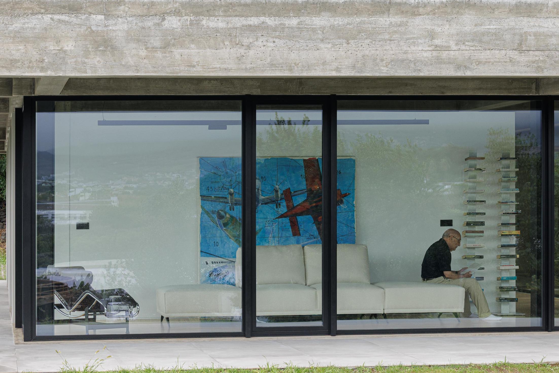 Sala de estar: Sofá branco, pintura e estante com livros. Grandes janelas com vista para o exterior. — Casa CR arquitetura SO Arquitetura & Design em São Miguel