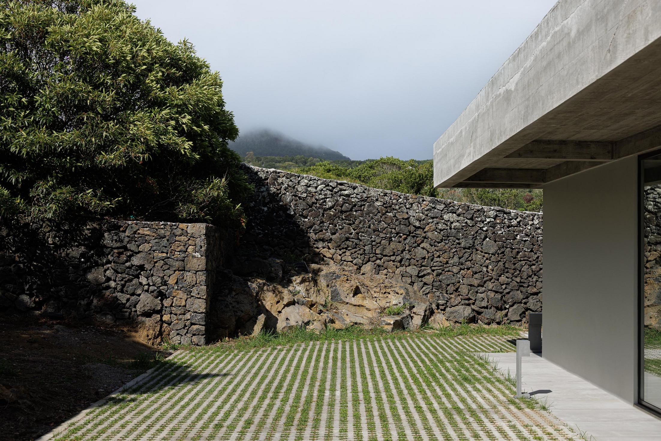Exterior: Muro de pedra vulcânica e fachada da casa com teto em betão aparente. — Casa CR arquitetura SO Arquitetura & Design em São Miguel