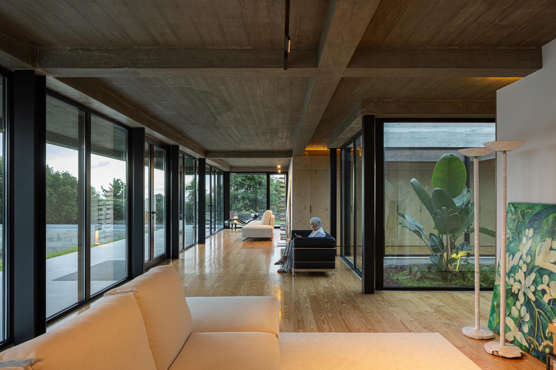 Sala de estar: Grandes janelas, sofá branco e cadeira preta. Pátio interior com vegetação. — Casa CR arquitetura SO Arquitetura & Design em São Miguel