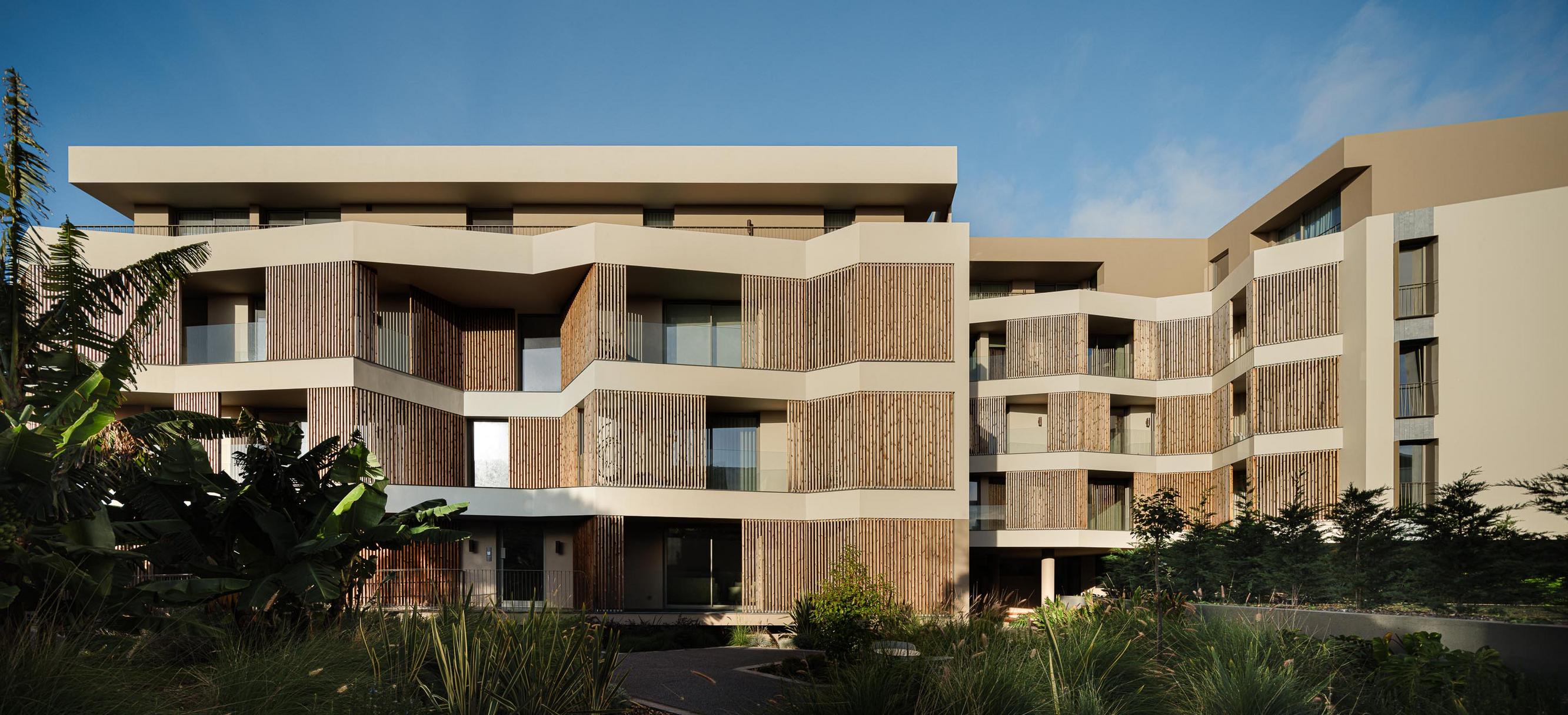 Fachada: Edifício com varandas protegidas por ripas de madeira, integrando-se na vegetação luxuriante. — ERNESTO DO CANTO - condomínio arquitetura BOX arquitectos em Ponta Delgada