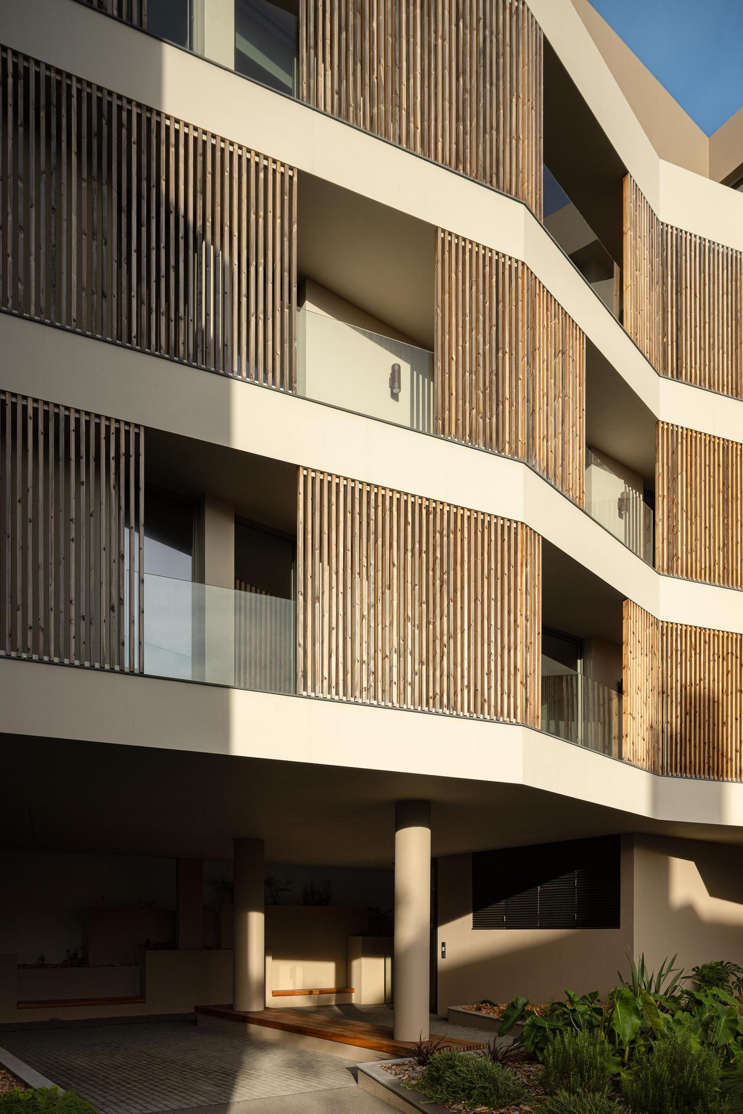 Fachada: Detalhe das varandas com ripas de madeira e vidro, com acesso ao jardim e pavimento em calçada. — ERNESTO DO CANTO - condomínio arquitetura BOX arquitectos em Ponta Delgada