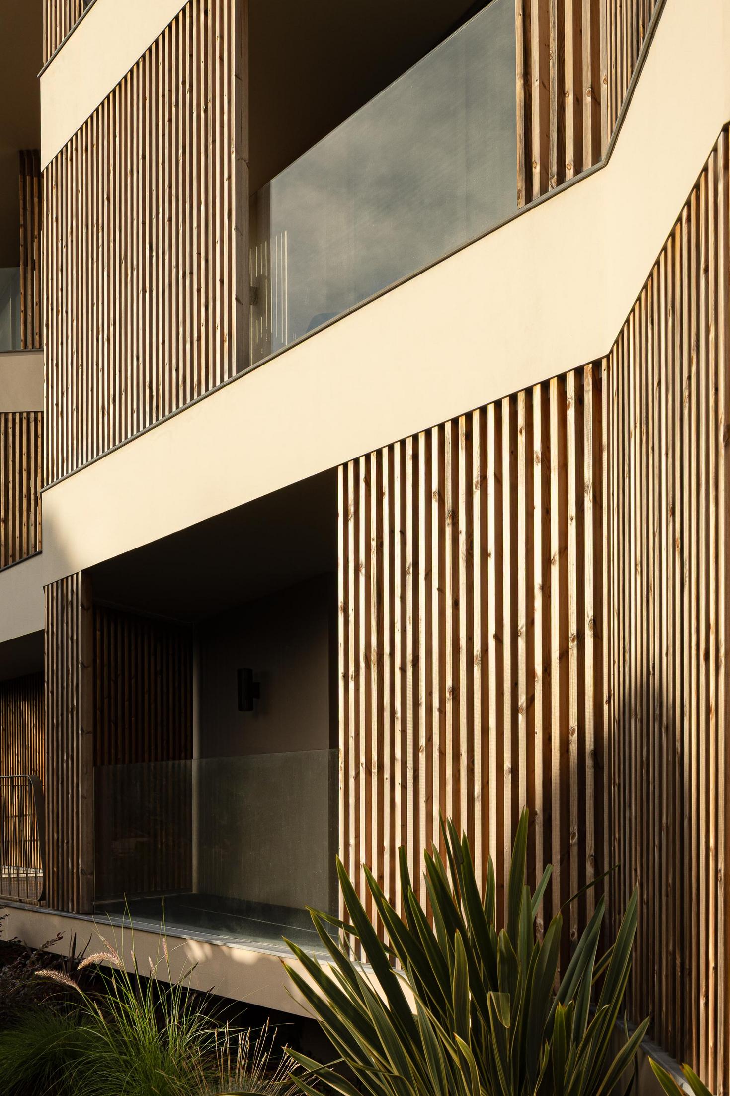 Fachada: Pormenor das ripas de madeira verticais e varanda com vidro, com vegetação rasteira. — ERNESTO DO CANTO - condomínio arquitetura BOX arquitectos em Ponta Delgada