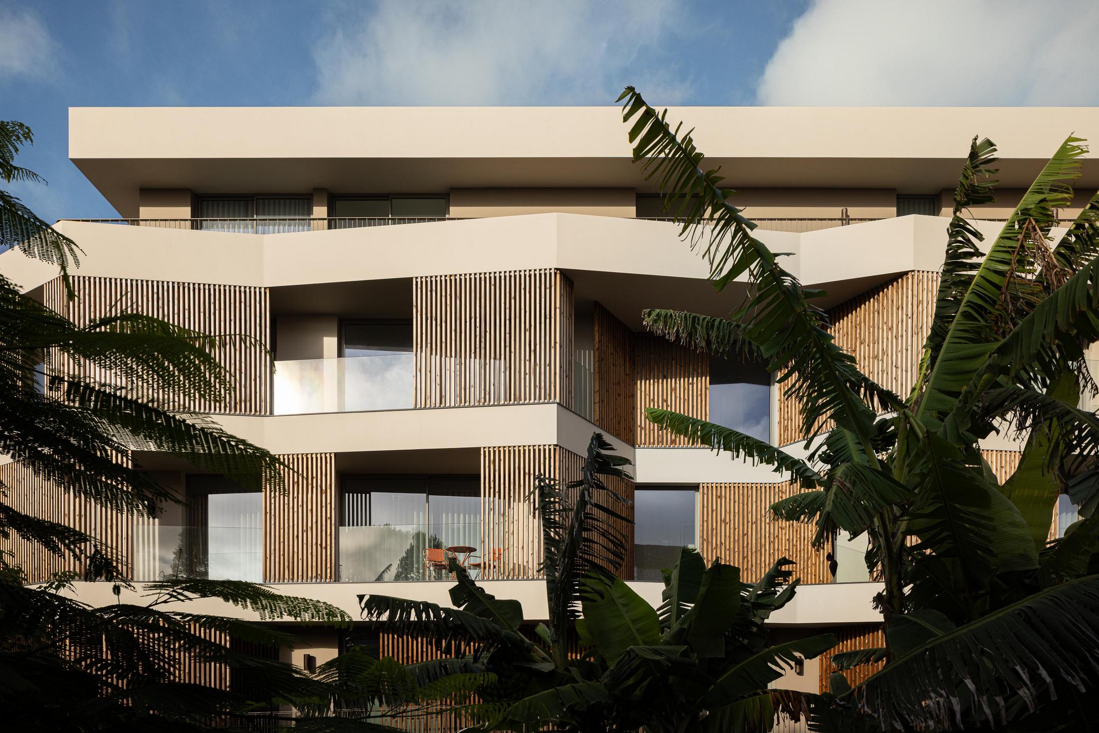 Fachada: Vista angular do edifício com ripas de madeira nas varandas e vegetação tropical. — ERNESTO DO CANTO - condomínio arquitetura BOX arquitectos em Ponta Delgada