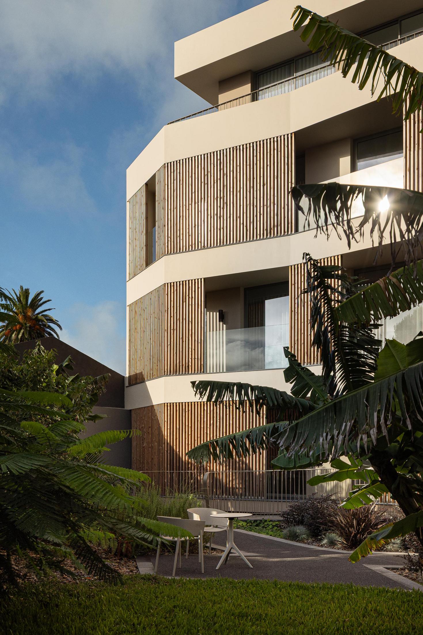 Fachada: Edifício com varandas em ripas de madeira, integrado num jardim tropical com palmeiras. — ERNESTO DO CANTO - condomínio arquitetura BOX arquitectos em Ponta Delgada