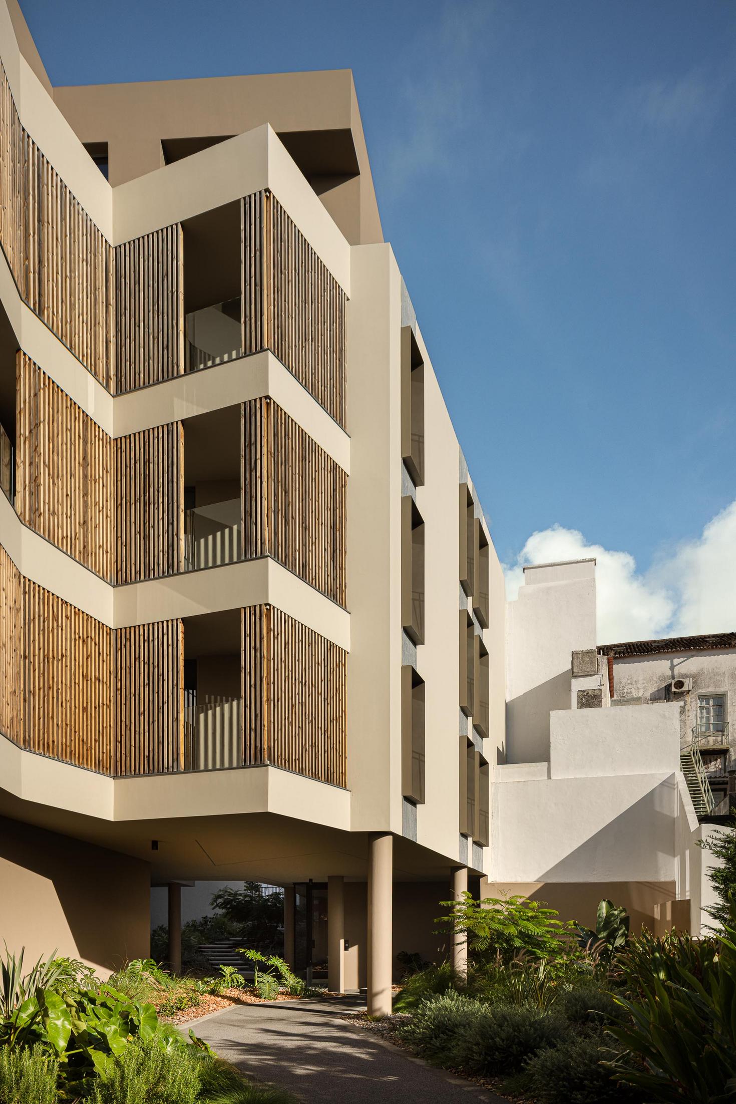 Fachada: Perspectiva do edifício com varandas em ripas de madeira e jardim com vegetação densa. — ERNESTO DO CANTO - condomínio arquitetura BOX arquitectos em Ponta Delgada