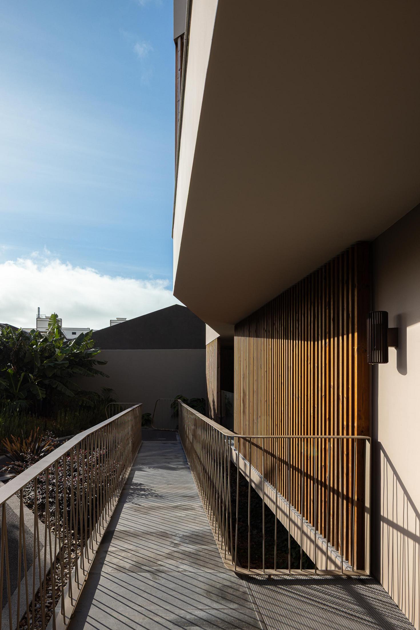 Rampa de acesso: Corredor exterior com ripas de madeira, candeeiro de parede e grade metálica. — ERNESTO DO CANTO - condomínio arquitetura BOX arquitectos em Ponta Delgada