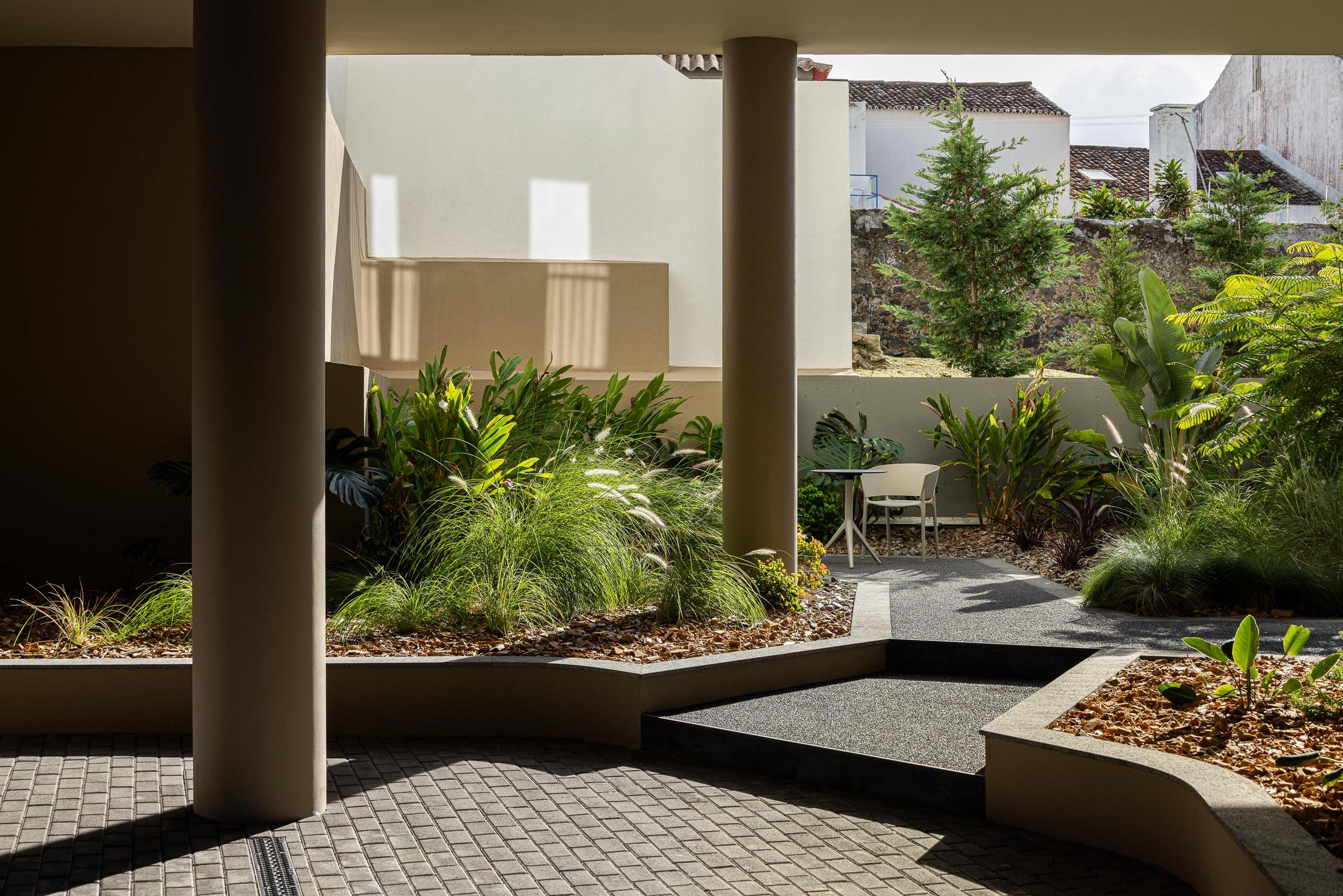 Pátio interior: Espaço ajardinado com vegetação exuberante, mesas e cadeiras, sob colunas de suporte. — ERNESTO DO CANTO - condomínio arquitetura BOX arquitectos em Ponta Delgada