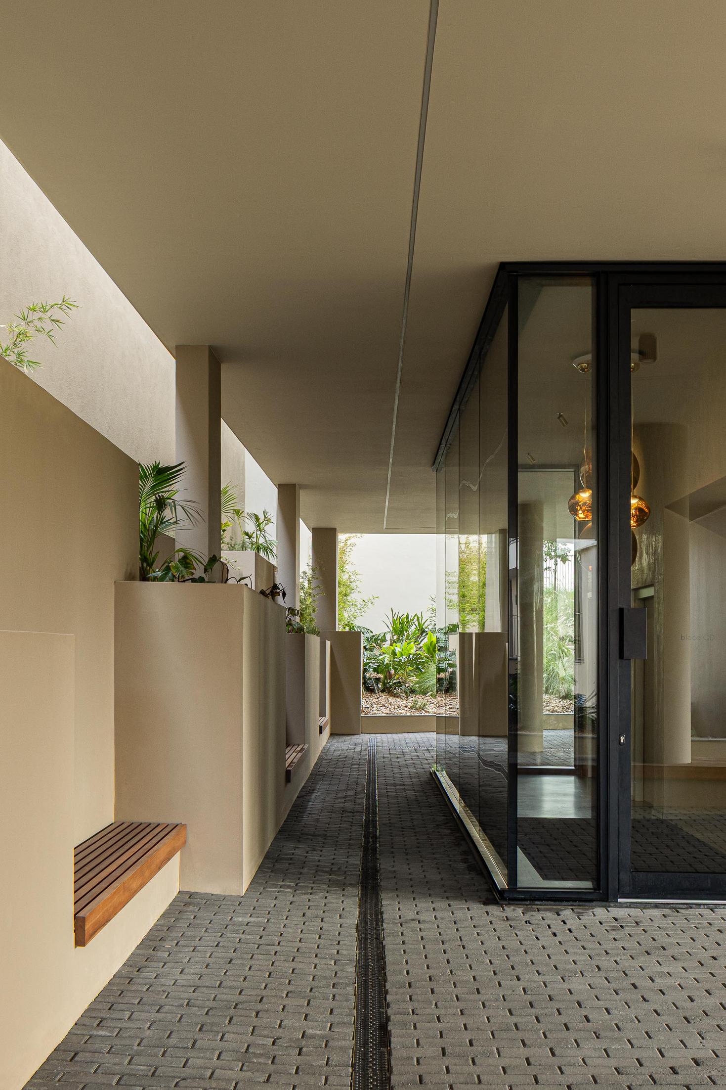 Corredor de acesso: Espaço coberto com pavimento em calçada, bancos e candeeiros suspensos. — ERNESTO DO CANTO - condomínio arquitetura BOX arquitectos em Ponta Delgada