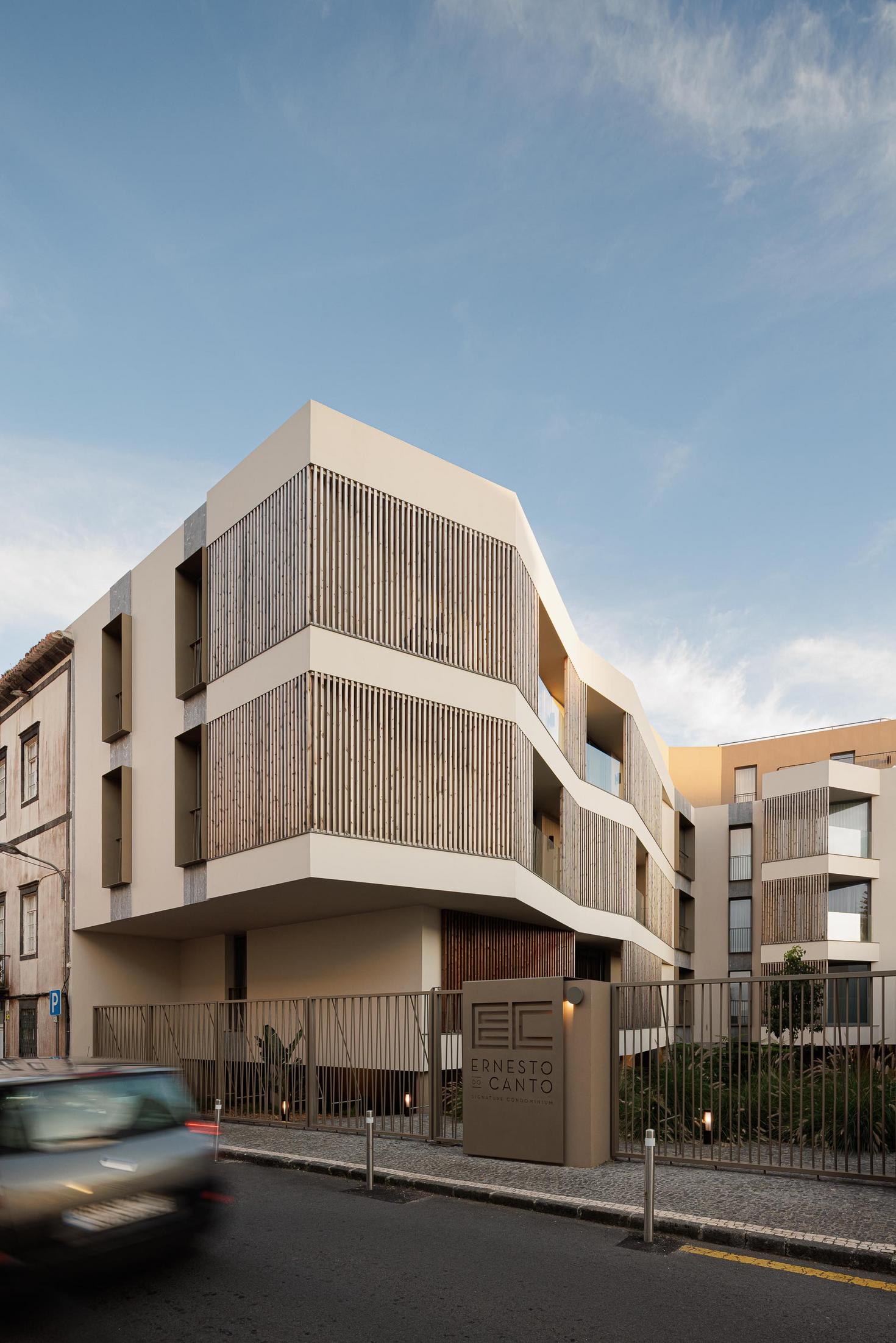Fachada: Vista do edifício com ripas de madeira, grade de acesso e vegetação. — ERNESTO DO CANTO - condomínio arquitetura BOX arquitectos em Ponta Delgada