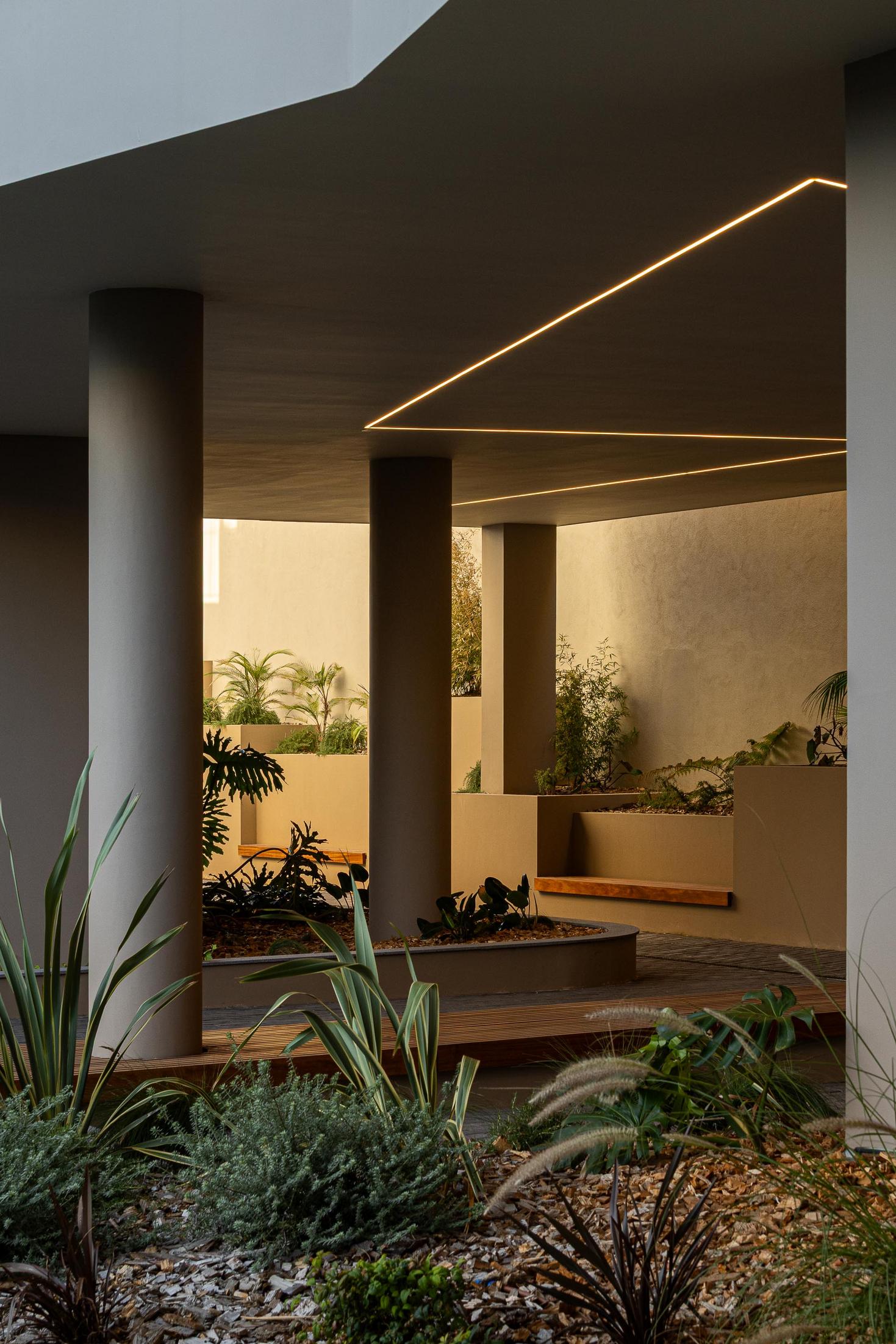Pátio interior: Espaço coberto com colunas, iluminação linear e vegetação tropical. — ERNESTO DO CANTO - condomínio arquitetura BOX arquitectos em Ponta Delgada