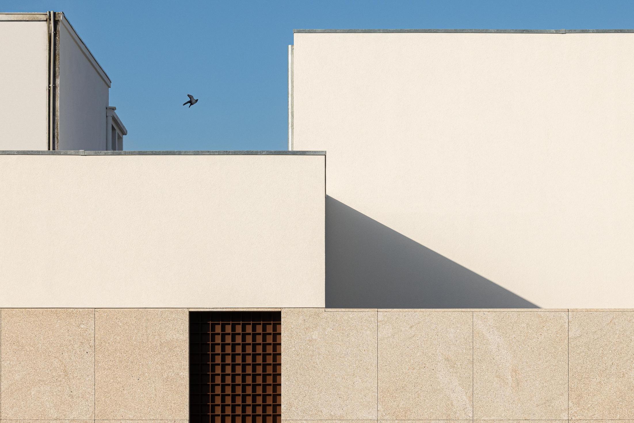 Exterior: Fachada com volumes geométricos em branco e granito, com grelha metálica. — Casa Mortuária São Martinho do Campo arquitetura Multiprojectus em Santo Tirso