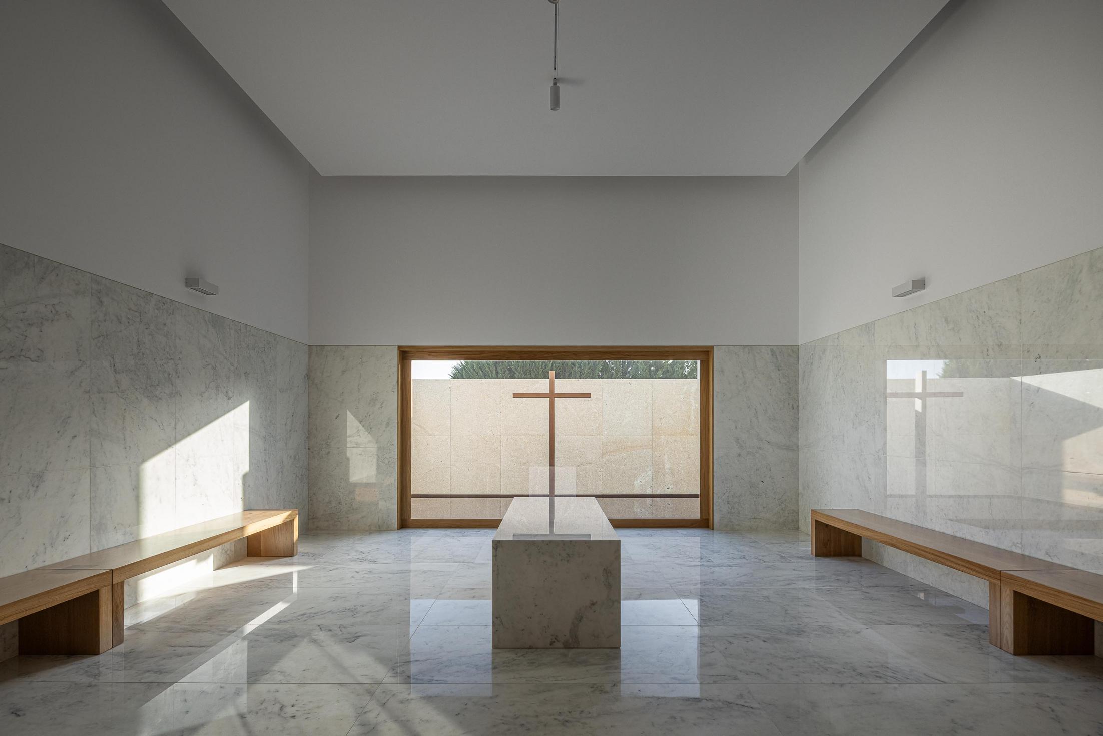 Capela: Interior minimalista com pavimento e paredes em mármore, bancos de madeira e cruz ao fundo. — Casa Mortuária São Martinho do Campo arquitetura Multiprojectus em Santo Tirso
