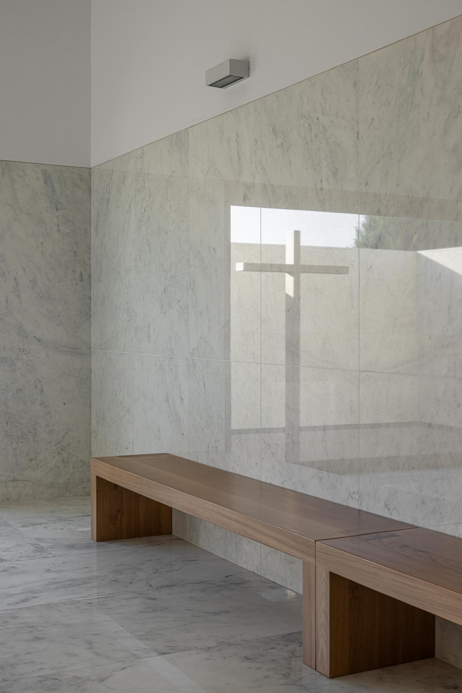 Capela: Banco de madeira e parede revestida a mármore, com reflexo da cruz na janela. — Casa Mortuária São Martinho do Campo arquitetura Multiprojectus em Santo Tirso