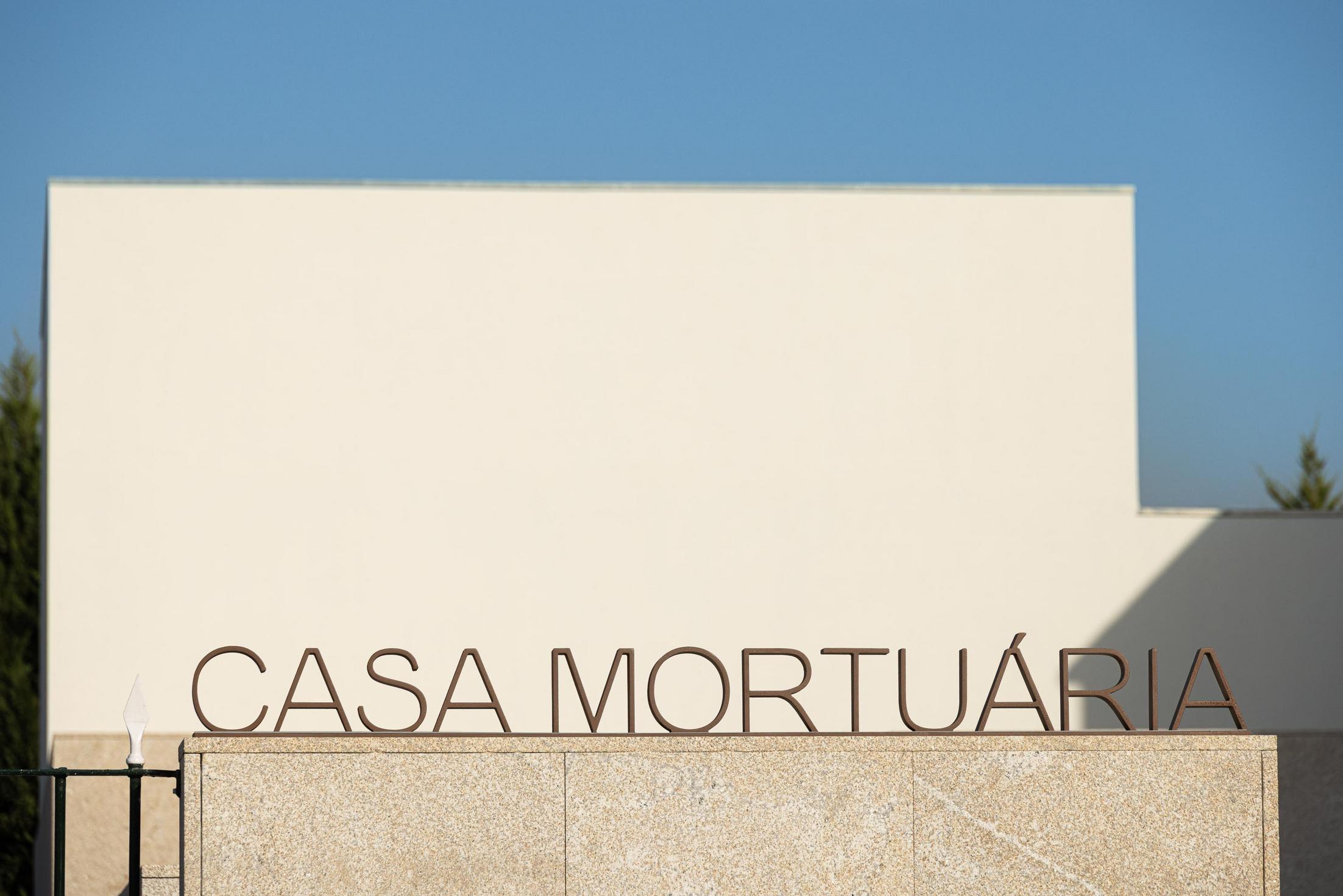 Exterior: Placa com o nome "Casa Mortuária" em letras metálicas, com fachada branca ao fundo. — Casa Mortuária São Martinho do Campo arquitetura Multiprojectus em Santo Tirso