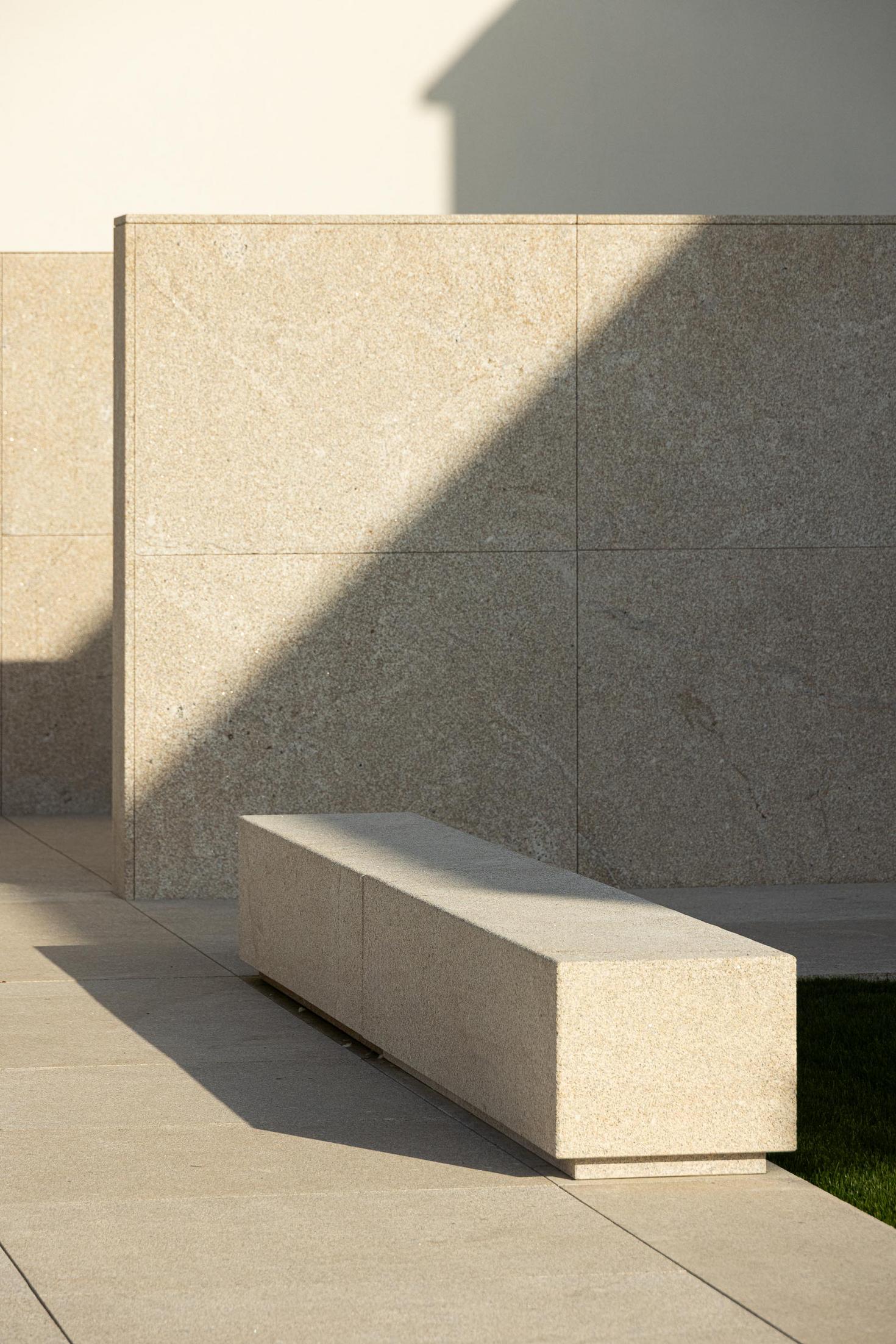 Exterior: Banco de granito no pátio, com relvado e paredes em granito. — Casa Mortuária São Martinho do Campo arquitetura Multiprojectus em Santo Tirso