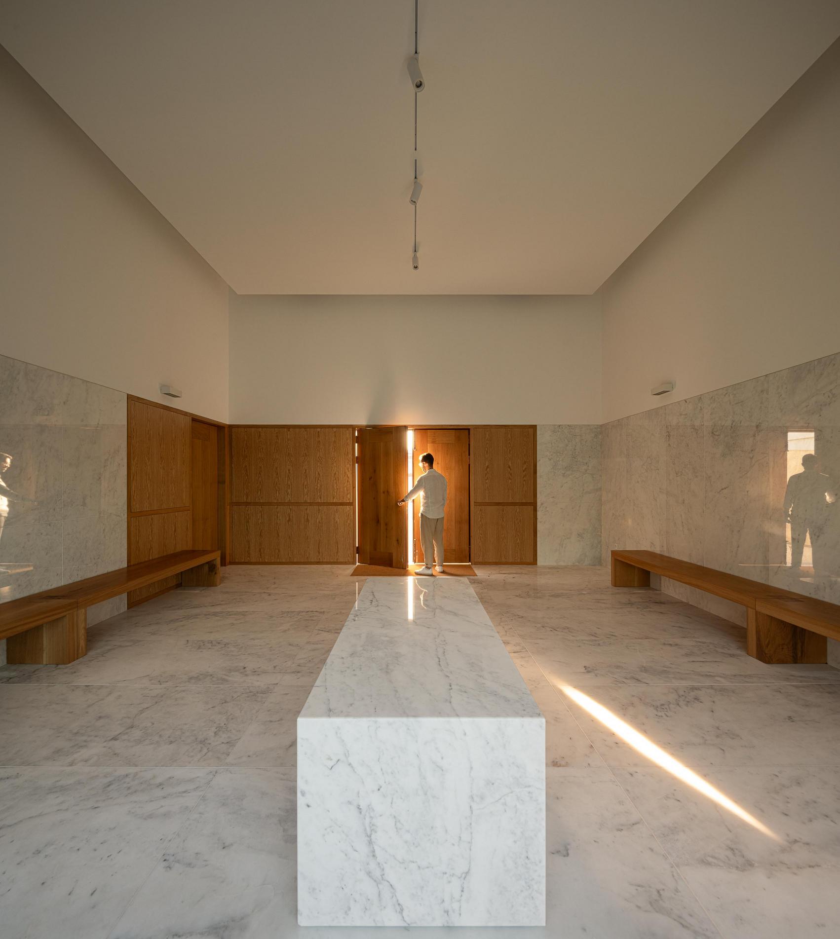 Capela: Interior com mesa em mármore, bancos de madeira e porta de madeira ao fundo. — Casa Mortuária São Martinho do Campo arquitetura Multiprojectus em Santo Tirso