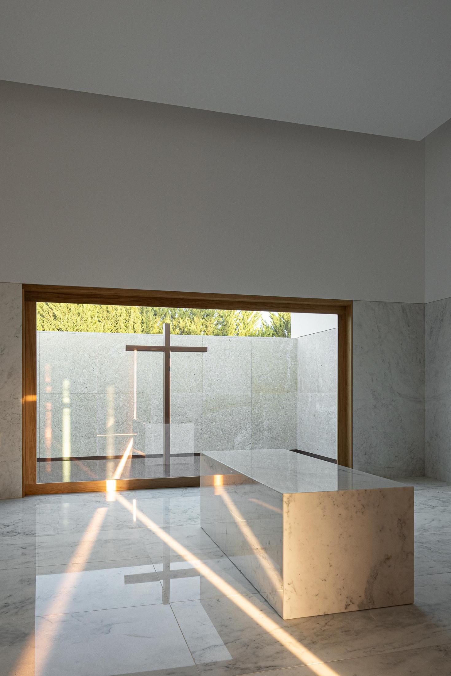 Capela: Mesa em mármore com reflexos da luz e vista para o pátio com cruz. — Casa Mortuária São Martinho do Campo arquitetura Multiprojectus em Santo Tirso