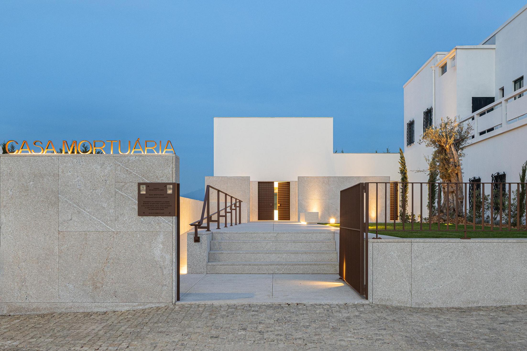 Exterior: Casa mortuária iluminada à noite, com placa identificativa e jardim. — Casa Mortuária São Martinho do Campo arquitetura Multiprojectus em Santo Tirso