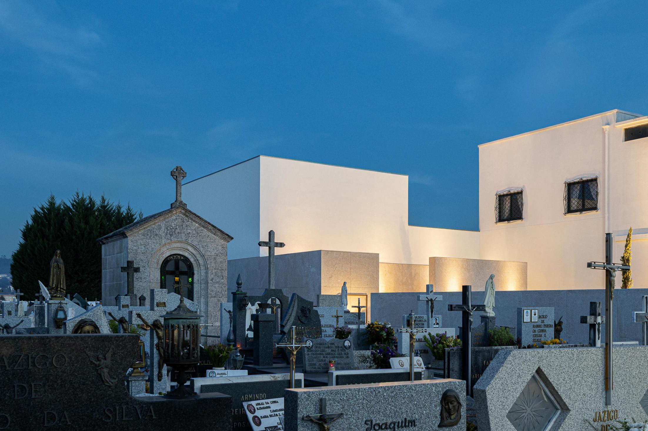 Exterior: Casa mortuária minimalista contrastando com as campas tradicionais iluminadas. — Casa Mortuária São Martinho do Campo arquitetura Multiprojectus em Santo Tirso