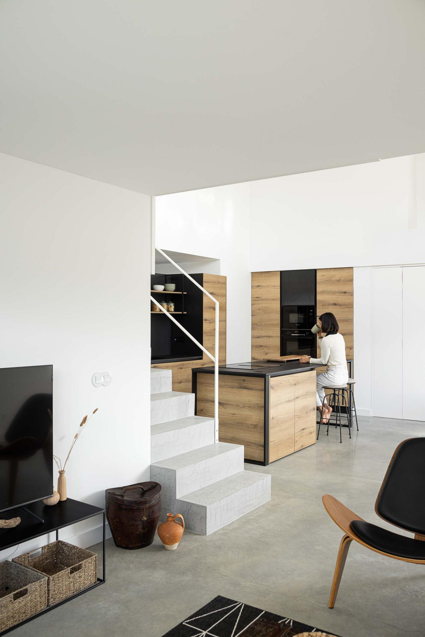 Sala de estar: Vista da sala de estar com escadas, cozinha em madeira e cadeiras pretas. — CASINHA DA MELROEIRA arquitetura Filipe Saraiva Arquitectos em Ourém