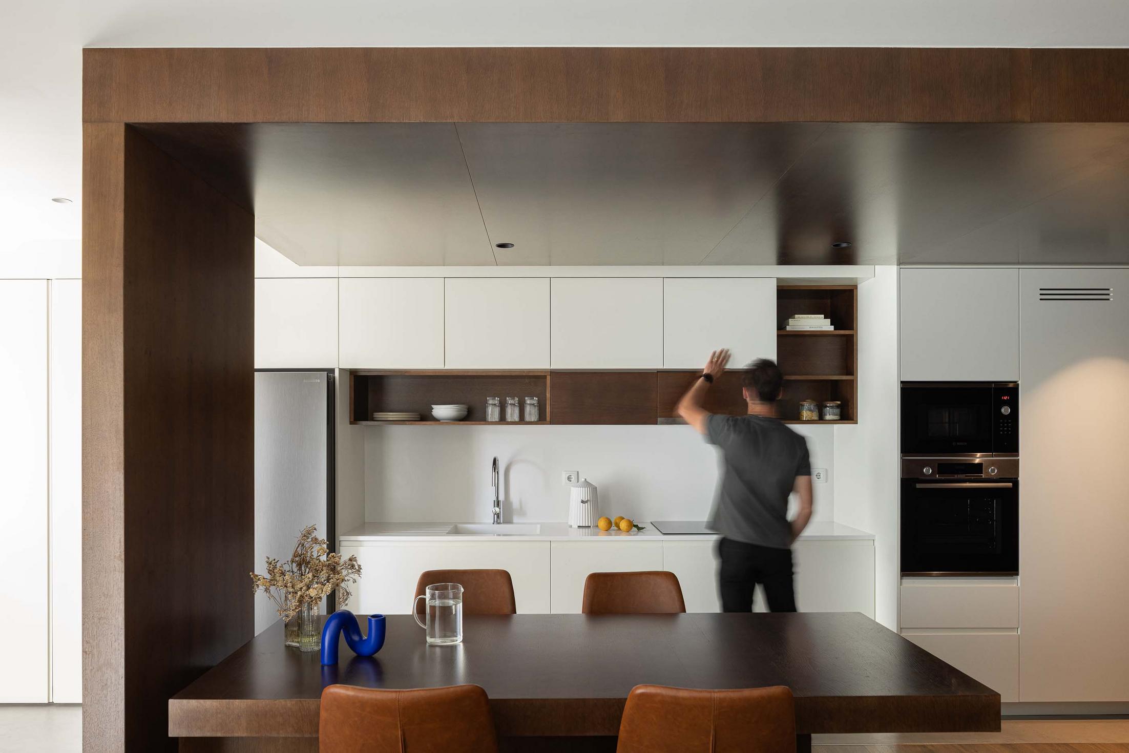 Cozinha/Sala de jantar: Homem a abrir armário branco. Mesa de madeira escura com cadeiras em pele castanha. Pavimento em madeira. — APARTAMENTO FP arquitetura ArquitectosRT em Braga