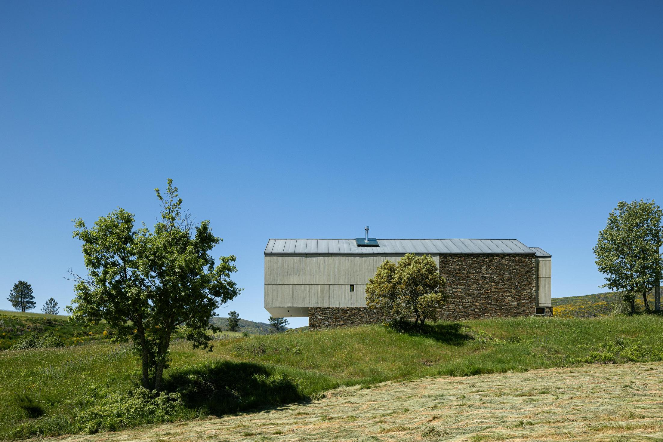 Exterior: Vista lateral da casa com revestimento em pedra e betão, integrada na paisagem campestre. — TerraSense Mountain Charm Retreat arquitetura DRK Atelier em Guarda