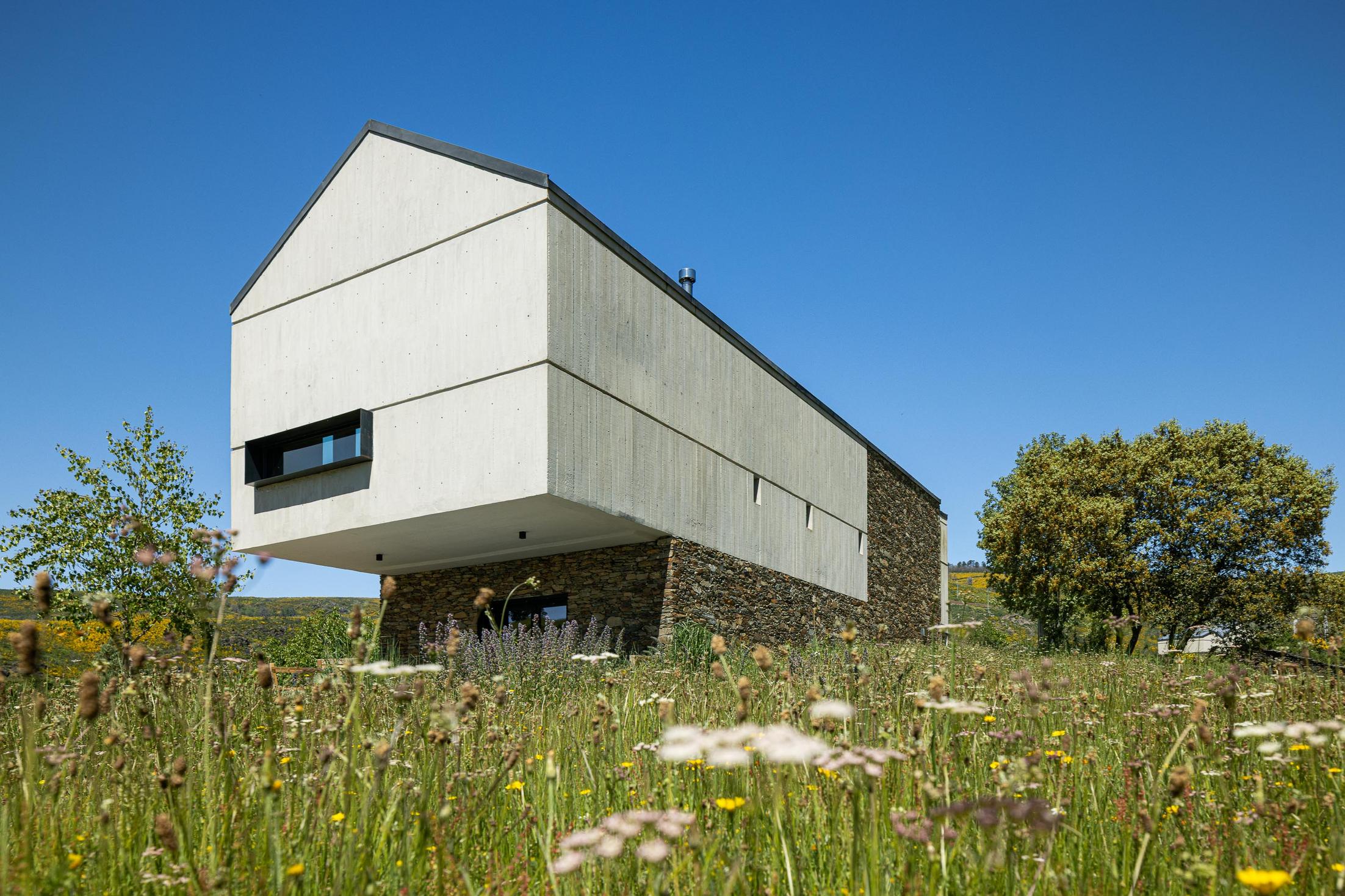 Exterior: Casa moderna com base em pedra e volume superior em betão, em campo florido. — TerraSense Mountain Charm Retreat arquitetura DRK Atelier em Guarda