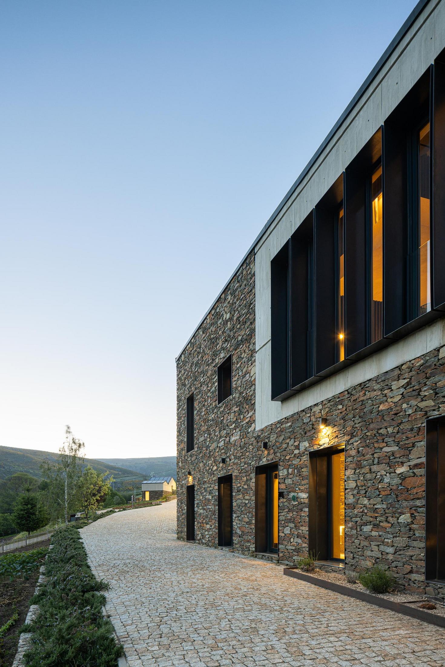Exterior: Casa com revestimento em pedra e betão, iluminada ao entardecer. — TerraSense Mountain Charm Retreat arquitetura DRK Atelier em Guarda