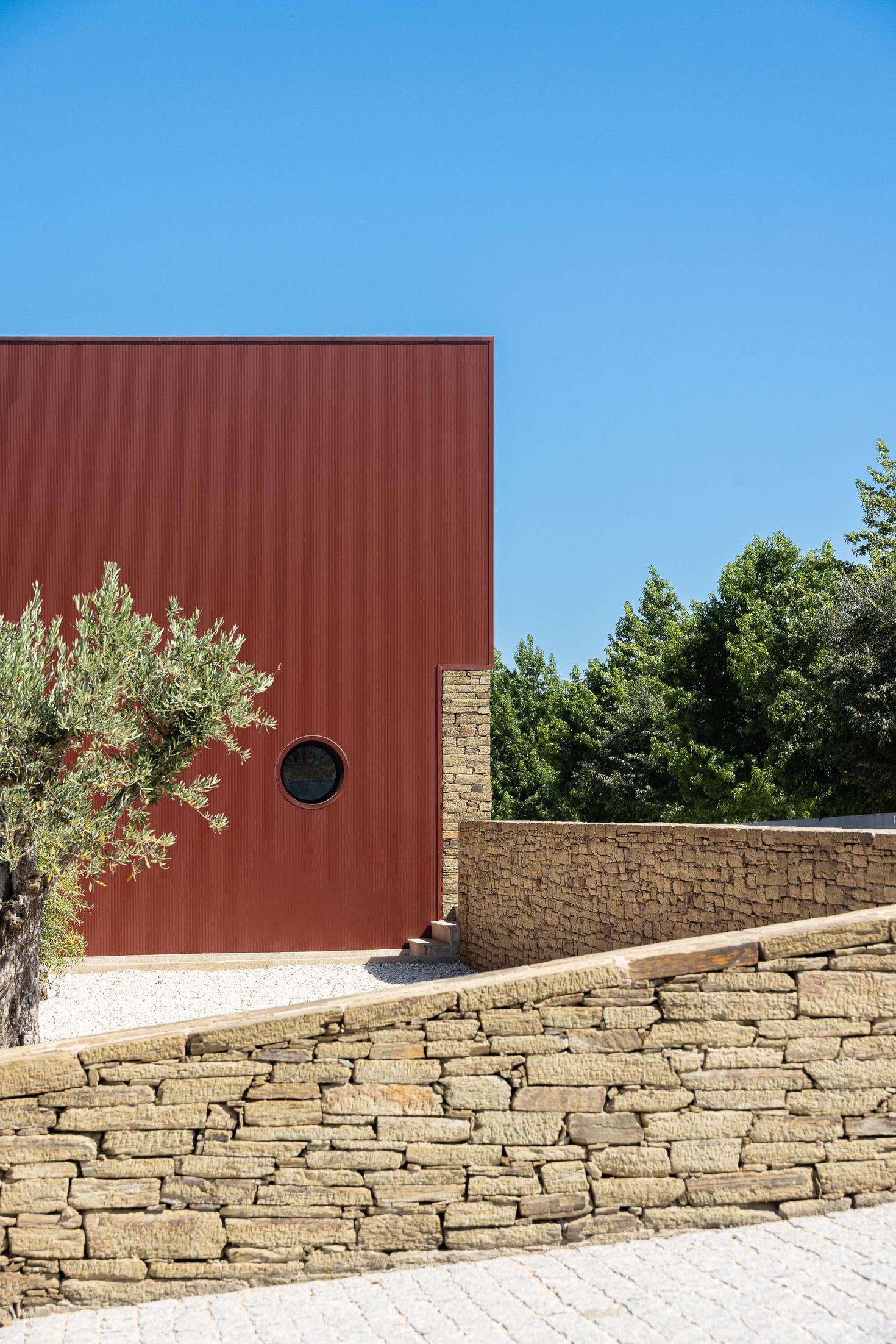 Exterior: Fachada com revestimento cor de vinho, janela circular e muro de pedra, com oliveira. — Adega Vinhos Borges arquitetura Em Paralelo em Vila Real