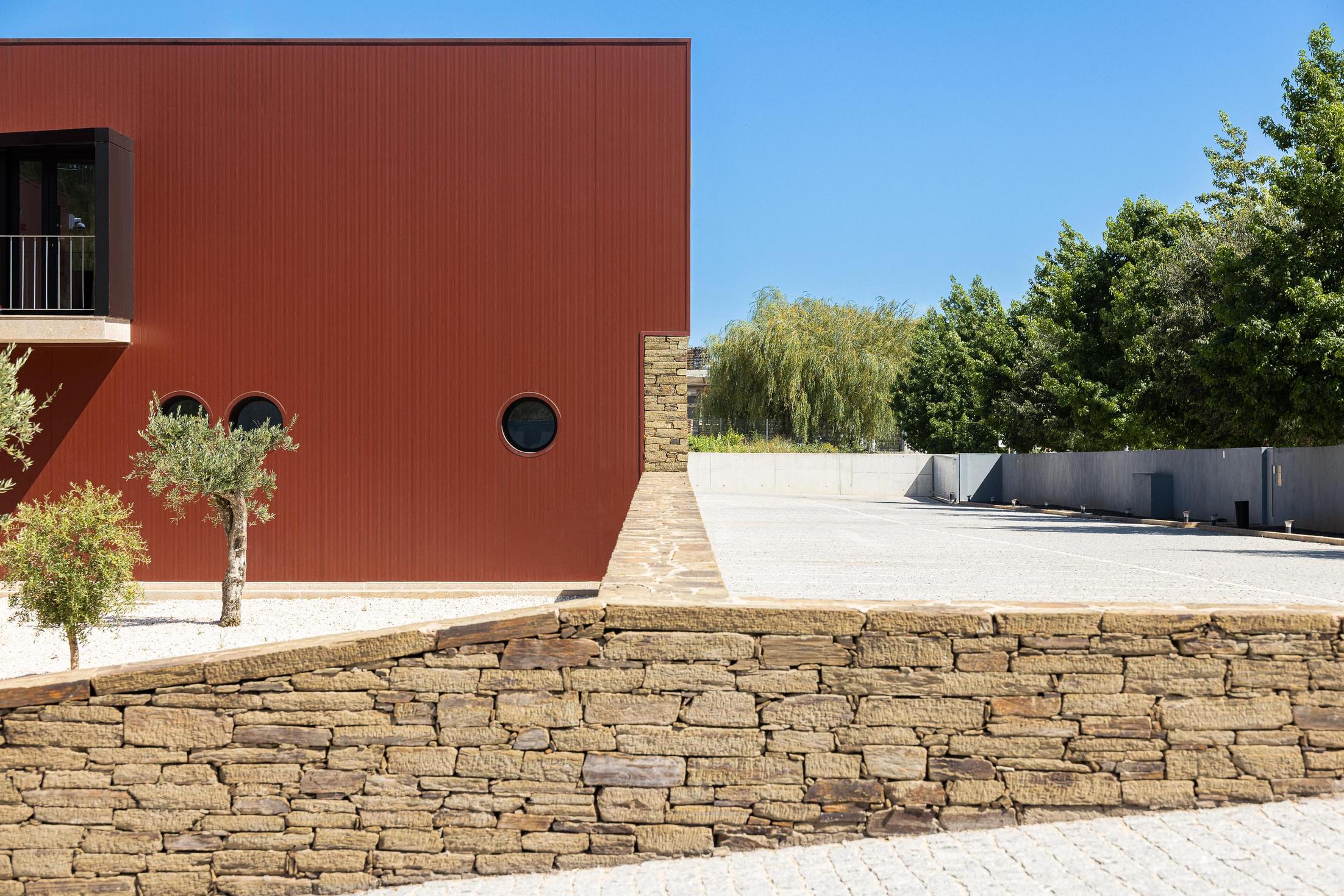 Exterior: Muro de pedra, fachada cor de vinho com janelas circulares e estacionamento. — Adega Vinhos Borges arquitetura Em Paralelo em Vila Real
