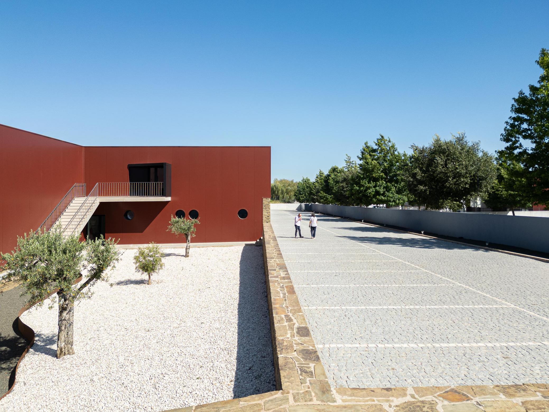 Exterior: Vista elevada do pátio com gravilha branca, oliveiras, estacionamento e fachada cor de vinho. — Adega Vinhos Borges arquitetura Em Paralelo em Vila Real