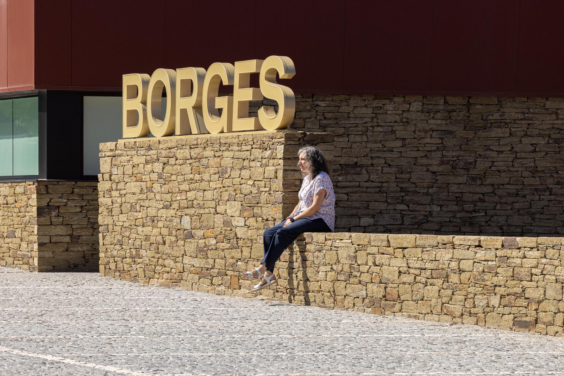 Exterior: Mulher sentada num muro de pedra com o nome "BORGES" em letras douradas ao fundo. — Adega Vinhos Borges arquitetura Em Paralelo em Vila Real