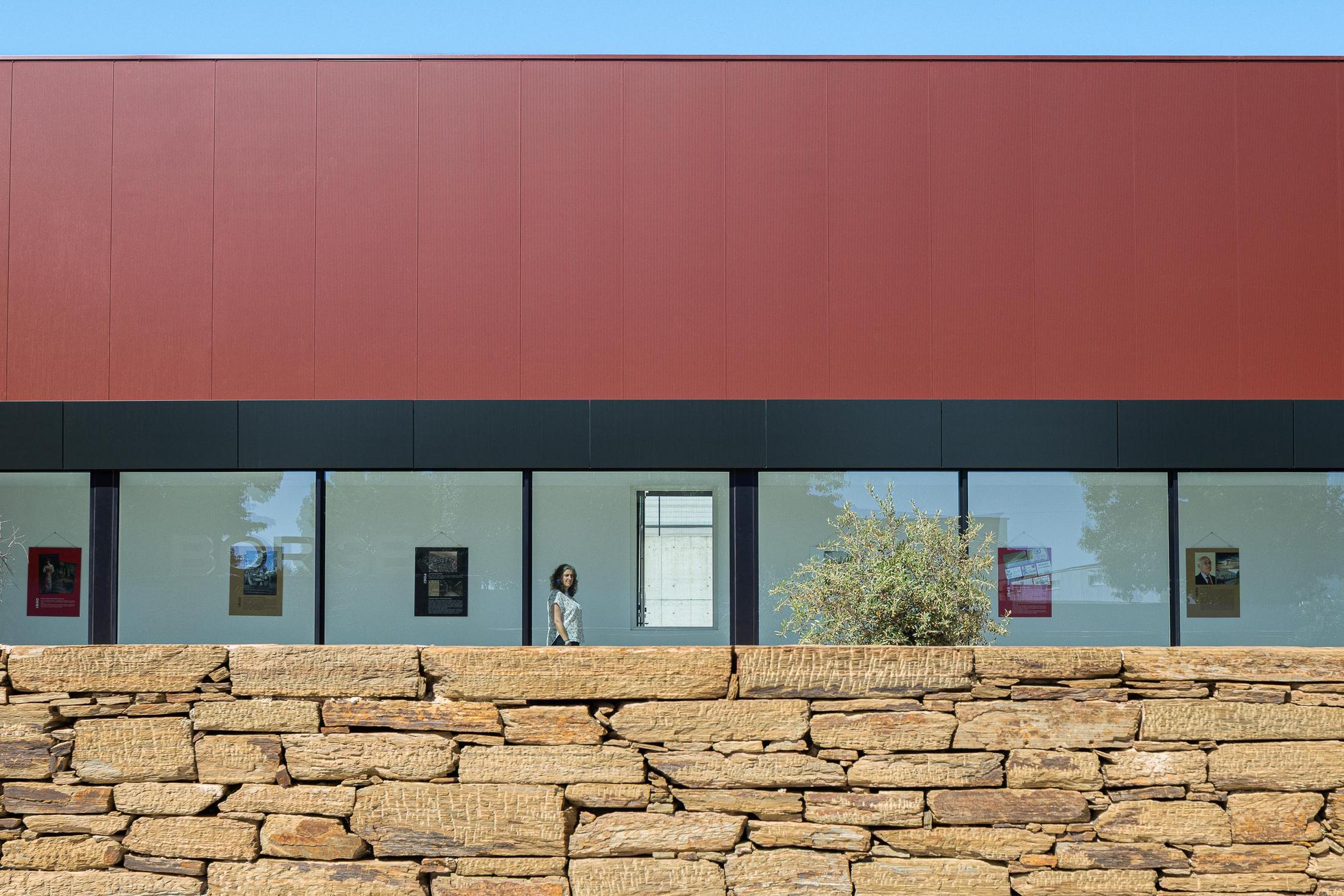 Galeria: Fachada envidraçada da galeria de arte com cartazes expostos e muro de pedra em primeiro plano. — Adega Vinhos Borges arquitetura Em Paralelo em Vila Real
