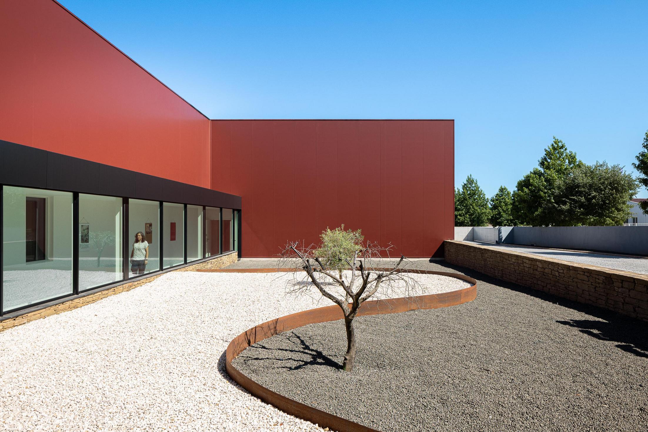 Jardim: Pátio exterior com gravilha branca e preta, oliveira e fachada cor de vinho com janelas. — Adega Vinhos Borges arquitetura Em Paralelo em Vila Real