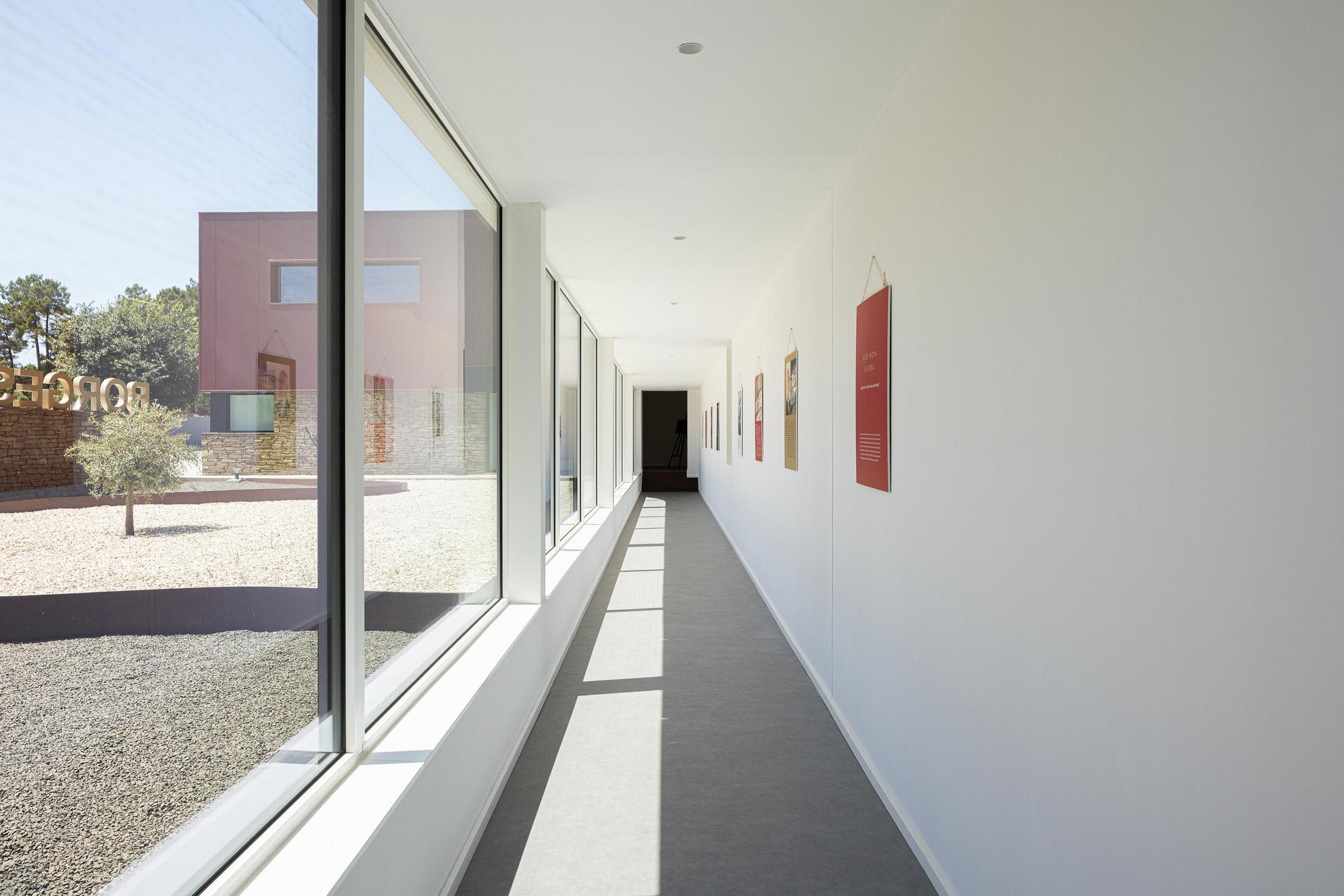Corredor: Corredor branco com pavimento cinzento, grandes janelas e cartazes nas paredes. — Adega Vinhos Borges arquitetura Em Paralelo em Vila Real