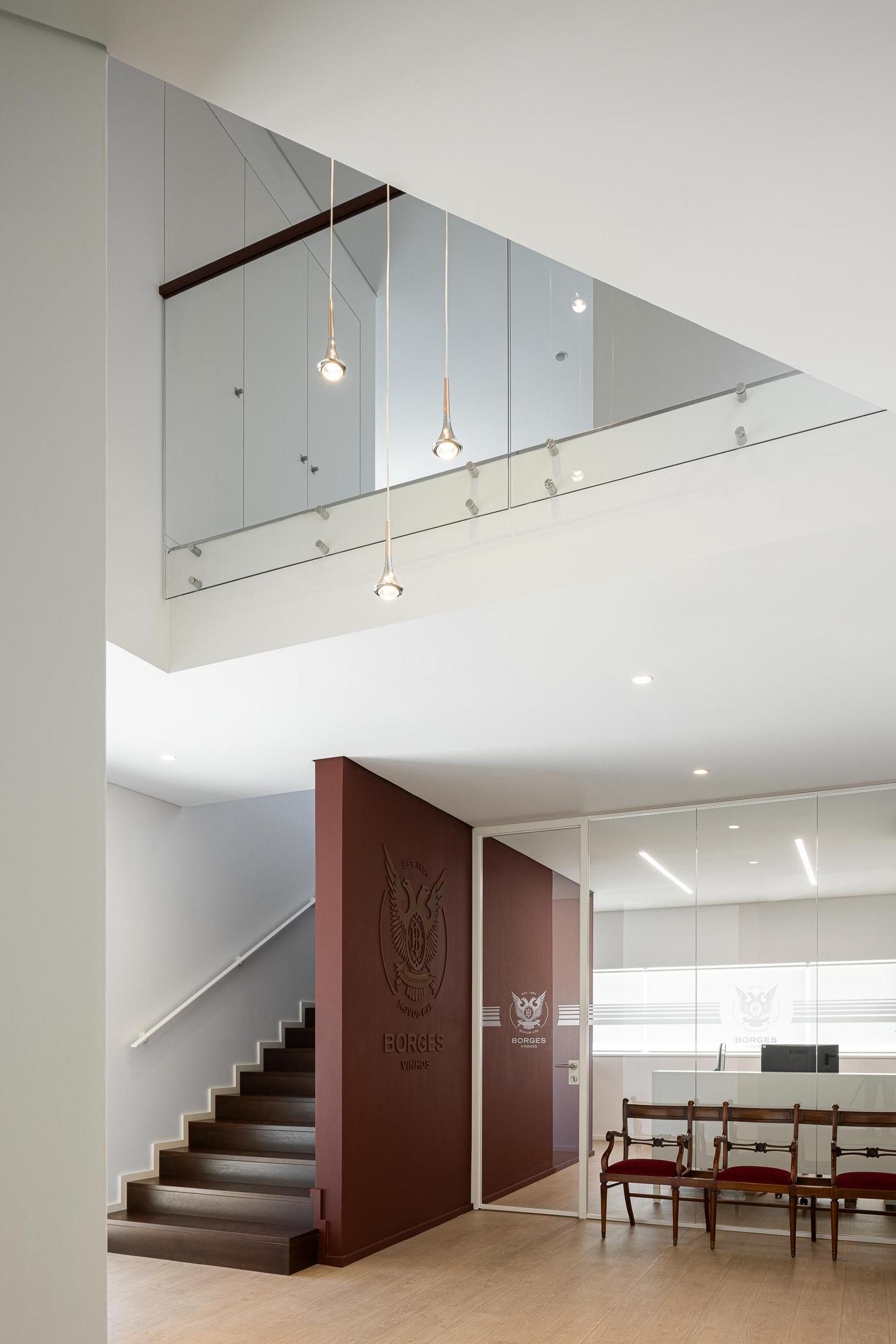 Recepção: Hall de entrada com escadaria em madeira, candeeiros pendentes e balcão de atendimento em vidro. — Adega Vinhos Borges arquitetura Em Paralelo em Vila Real