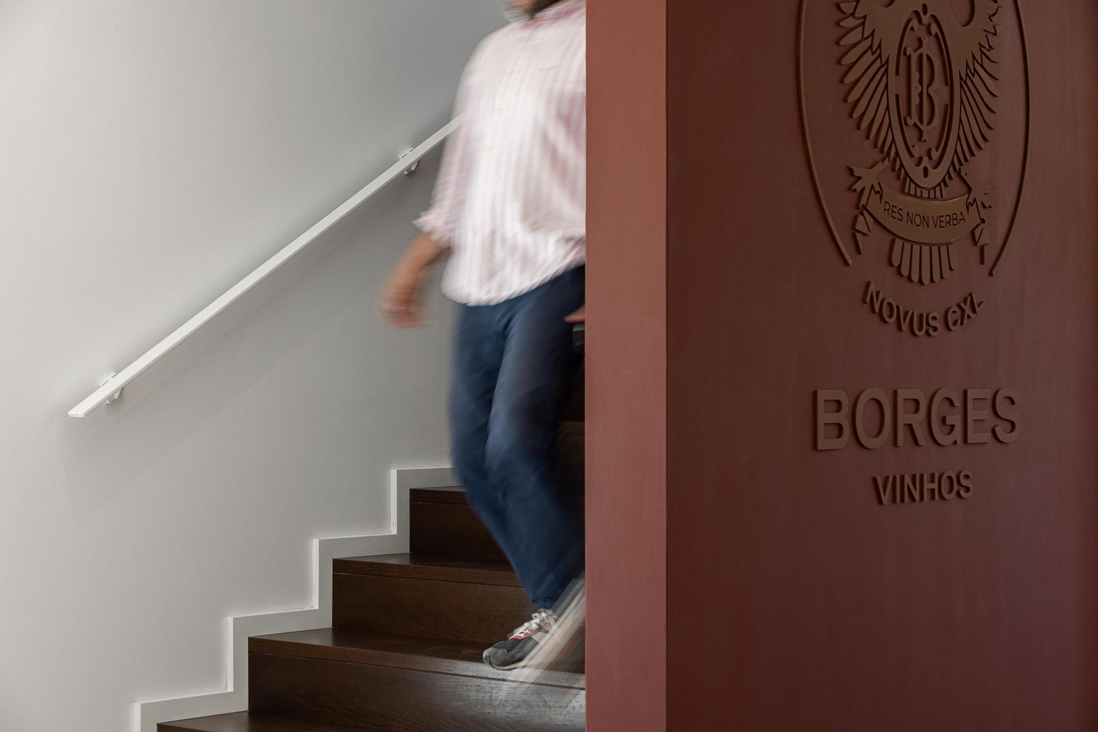Escadaria: Detalhe da escadaria em madeira com corrimão branco e parede cor de vinho com o brasão "BORGES". — Adega Vinhos Borges arquitetura Em Paralelo em Vila Real