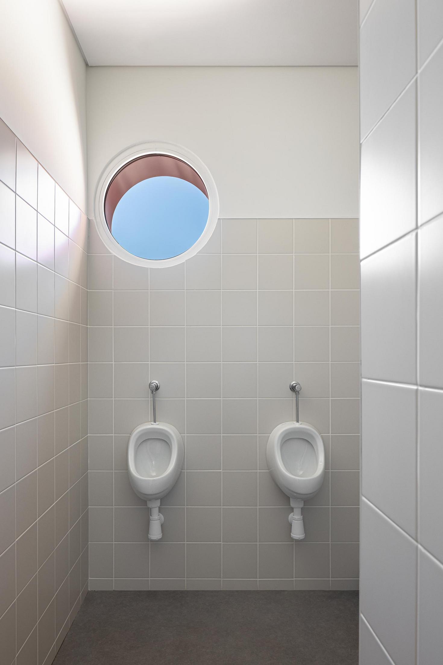 Casa de banho: Casa de banho masculina com dois urinóis, revestimento cerâmico cinzento e janela circular. — Adega Vinhos Borges arquitetura Em Paralelo em Vila Real