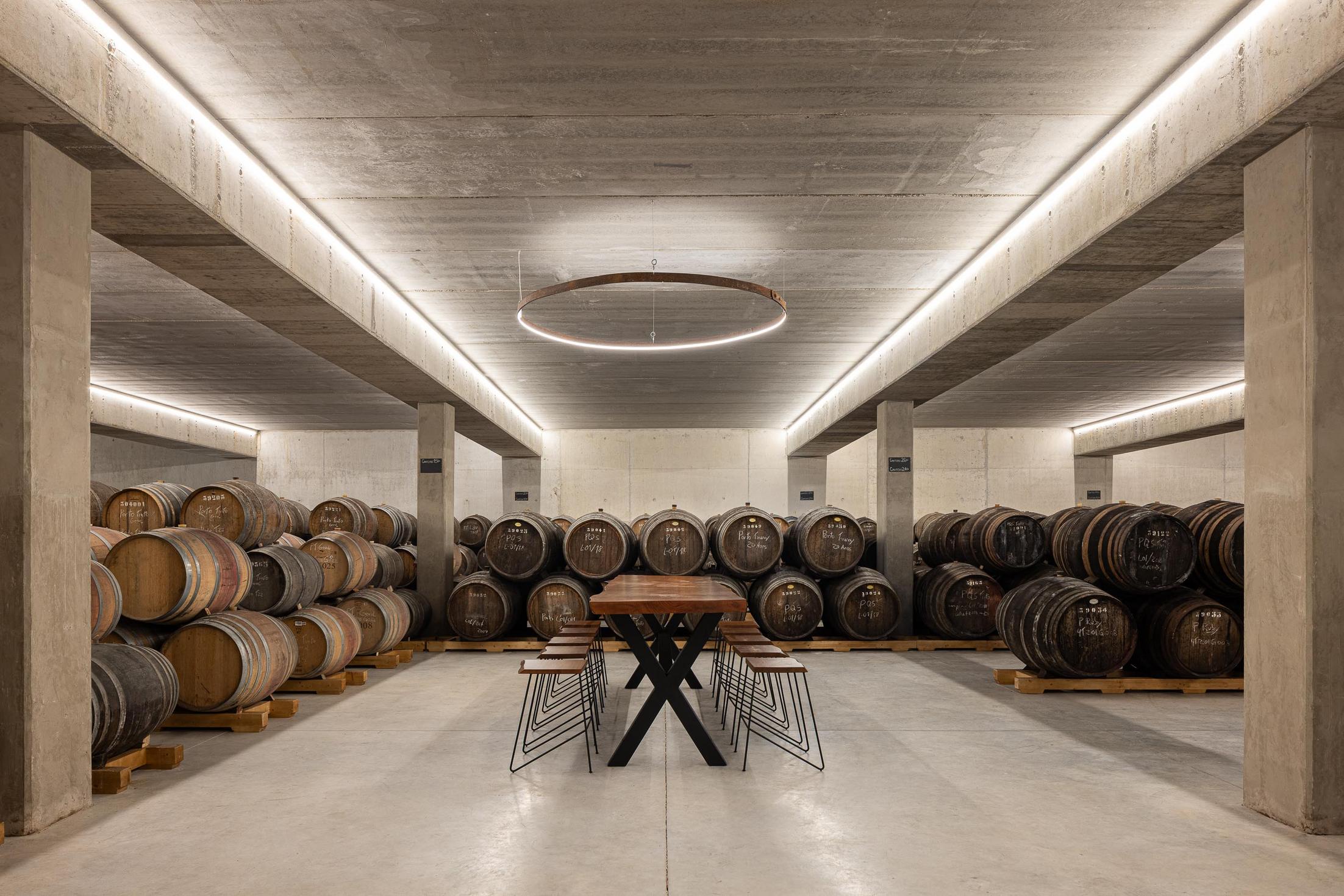 Cave: Cave com barricas de vinho, pavimento em betão e candeeiro circular suspenso. — Adega Vinhos Borges arquitetura Em Paralelo em Vila Real