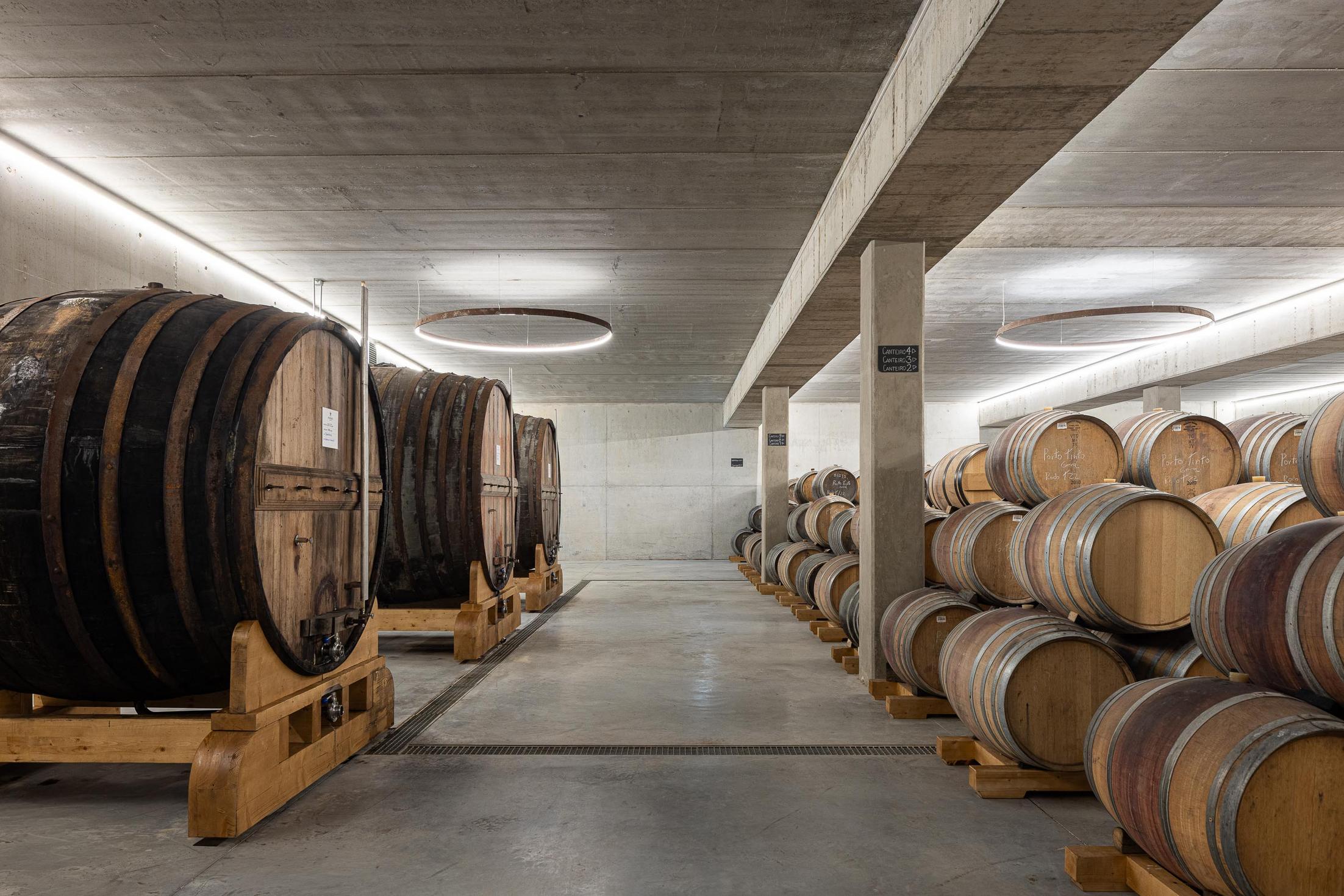 Cave: Cave com barricas de vinho alinhadas e candeeiro circular suspenso. — Adega Vinhos Borges arquitetura Em Paralelo em Vila Real