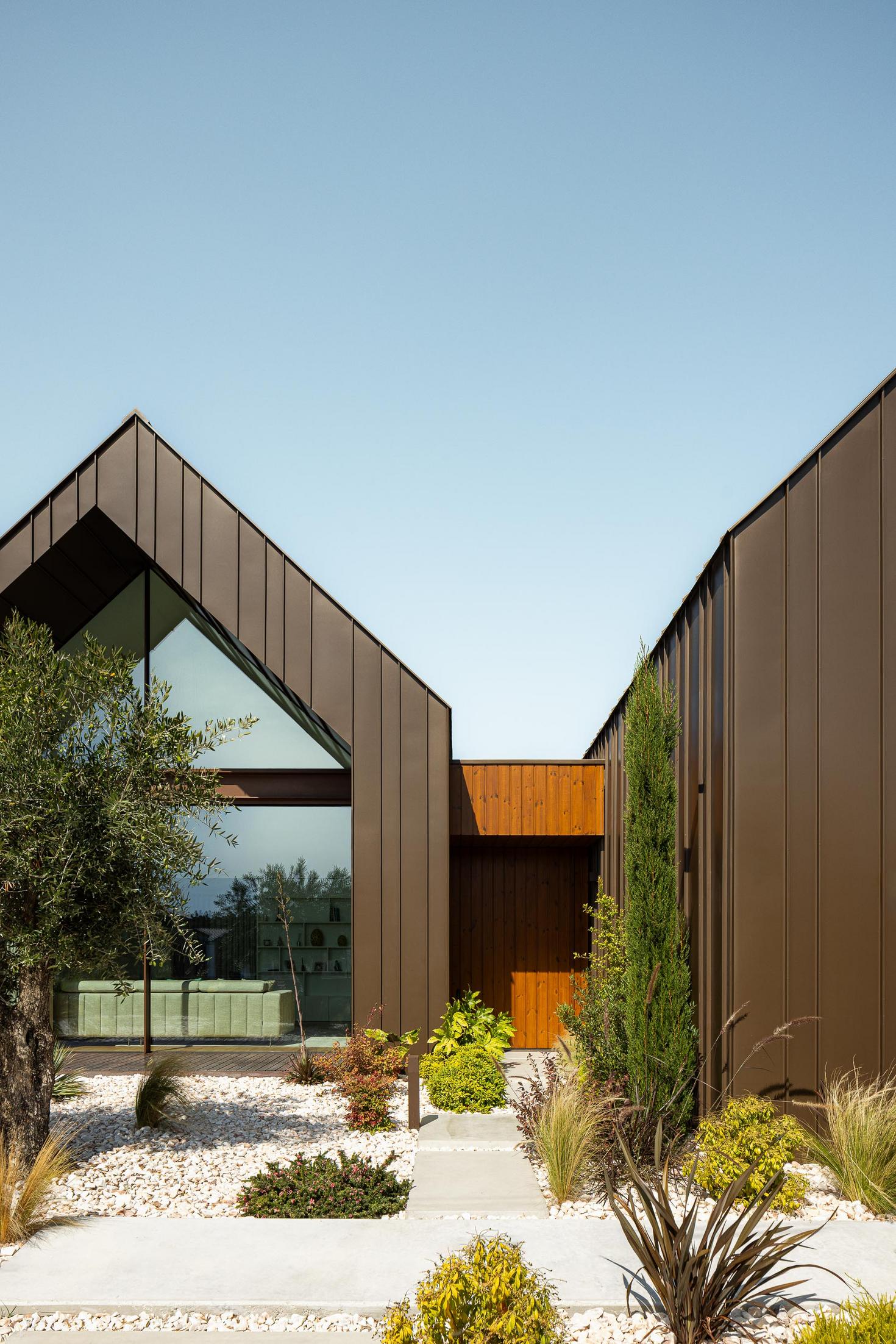 Entrada: Fachada com revestimento metálico castanho, janela triangular e caminho de pedra branca. — House SM arquitetura Mário Alves Arquitecto em Ílhavo