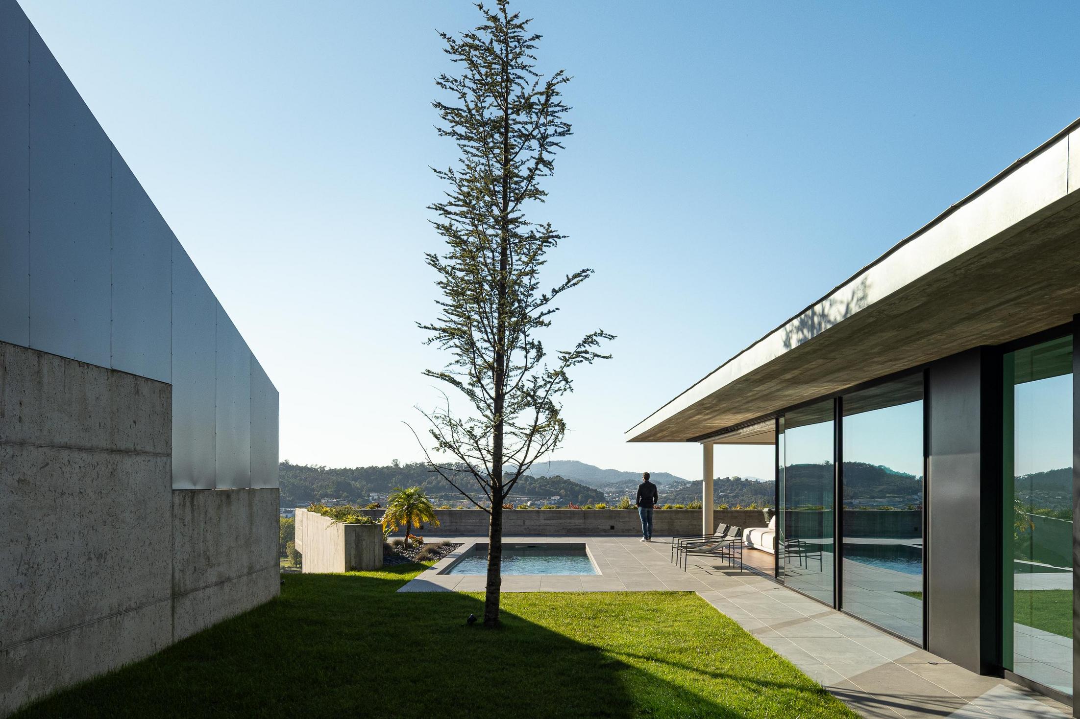 Exterior: Detalhe da fachada em betão aparente, com árvore jovem e vegetação rasteira. — CASA A arquitetura L2C Arquitetura em Braga