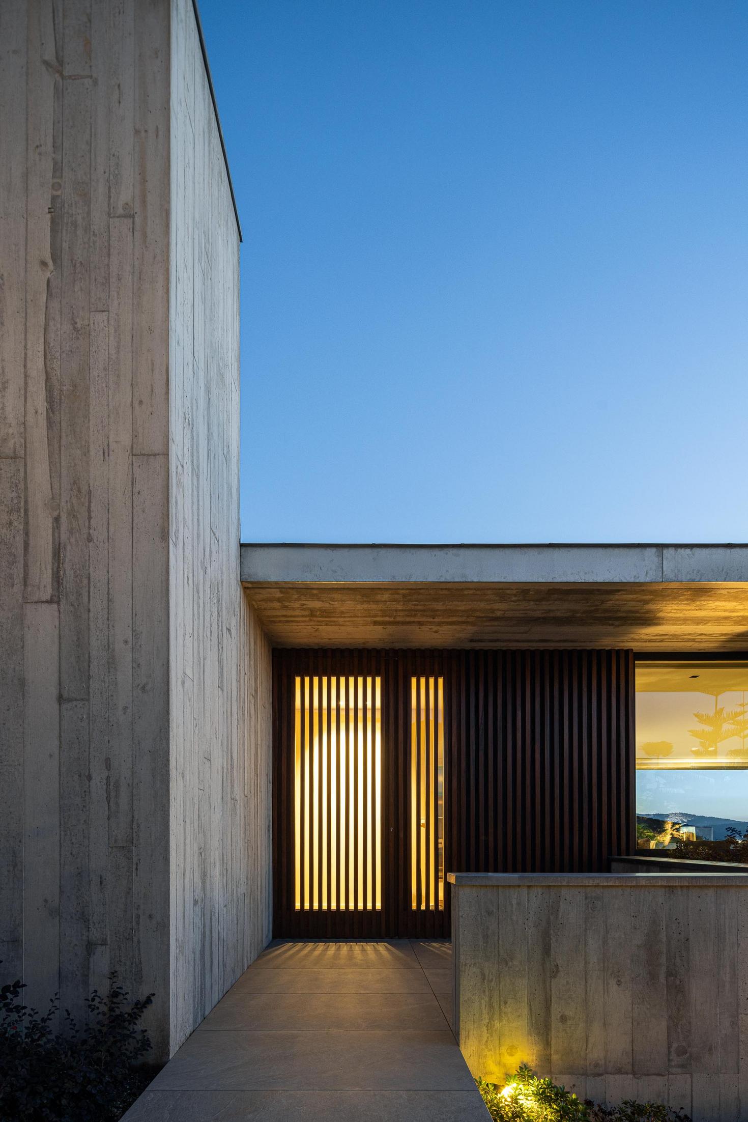 Garagem: Garagem com porta aberta e fachada em betão aparente iluminada ao anoitecer. — CASA A arquitetura L2C Arquitetura em Braga