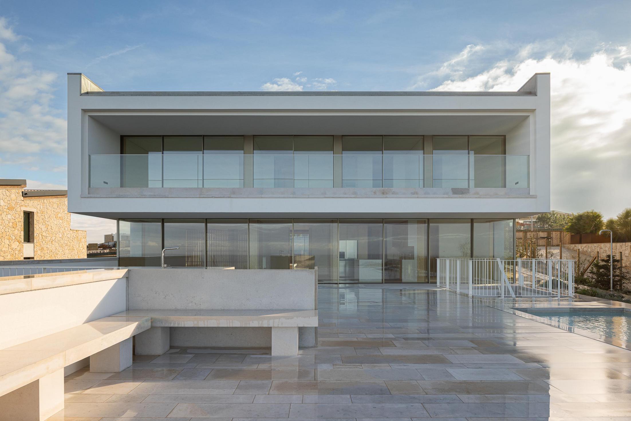 Fachada posterior: Casa minimalista com grandes janelas, terraço em pedra, piscina e banco em betão. — WEST CLIFFS D45 arquitetura ARX Portugal - Arquitectos em Óbidos