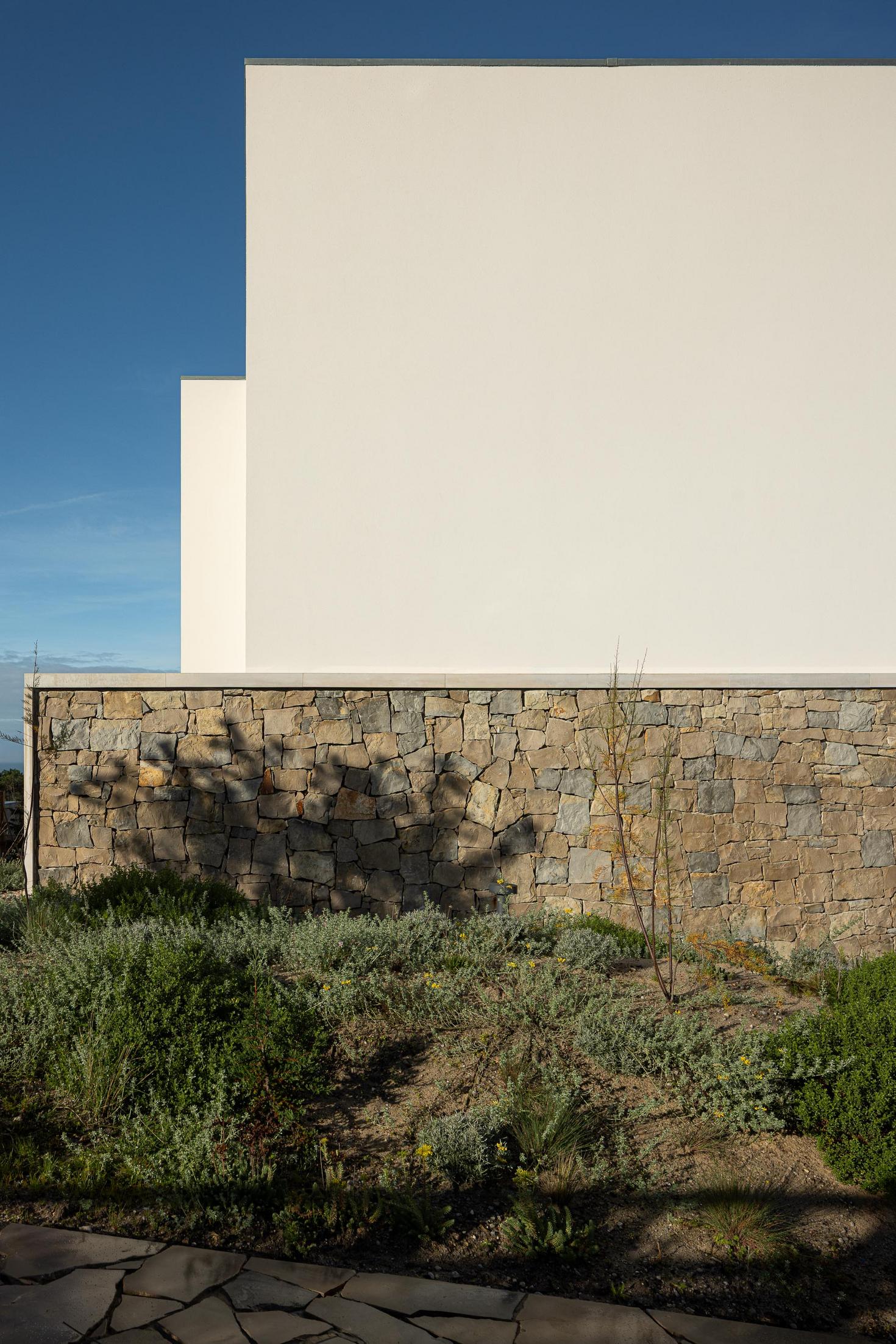 Fachada lateral: Detalhe da fachada com parede em pedra rústica e reboco branco, sob um céu azul. — WEST CLIFFS D45 arquitetura ARX Portugal - Arquitectos em Óbidos