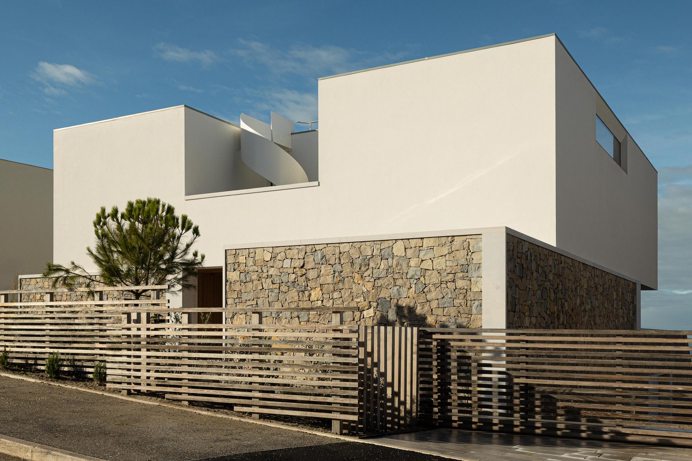 Fachada lateral: Perspectiva da casa com escada em espiral, pedra rústica e vedação de madeira. — WEST CLIFFS D45 arquitetura ARX Portugal - Arquitectos em Óbidos