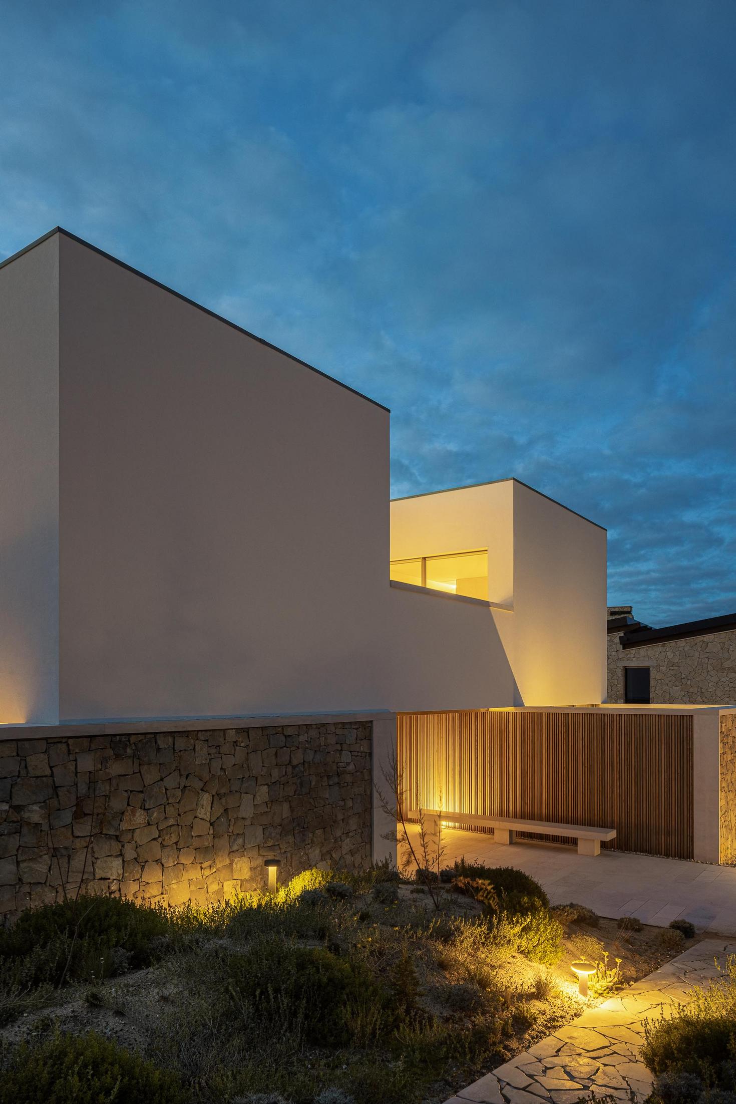 Fachada frontal: Vista noturna da fachada frontal com jardim, vedação em madeira e iluminação. — WEST CLIFFS D45 arquitetura ARX Portugal - Arquitectos em Óbidos